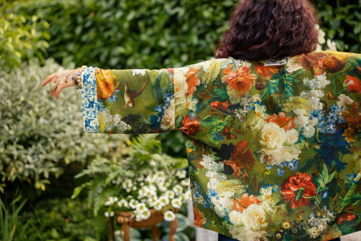 A person photographed from behind wearing a loose, long-sleeve floral jacket in an outdoor garden setting. The jacket features a dense multicolor floral and bird print with green, orange, white, and blue tones | Bliss by Cori