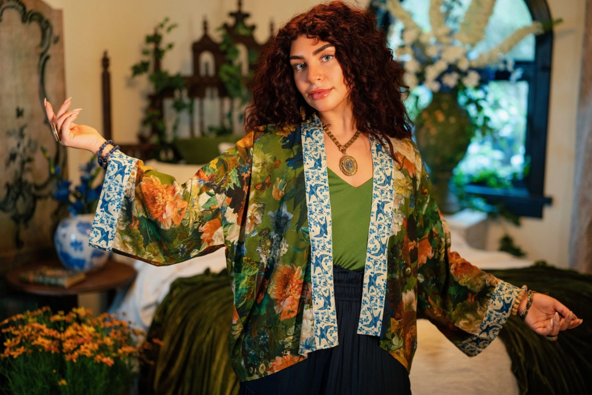 A woman with curly brown hair models a bohemian-style floral kimono jacket with blue patterned trim over a green top, standing in a decorated room with plants and vintage decor | Bliss by Cori