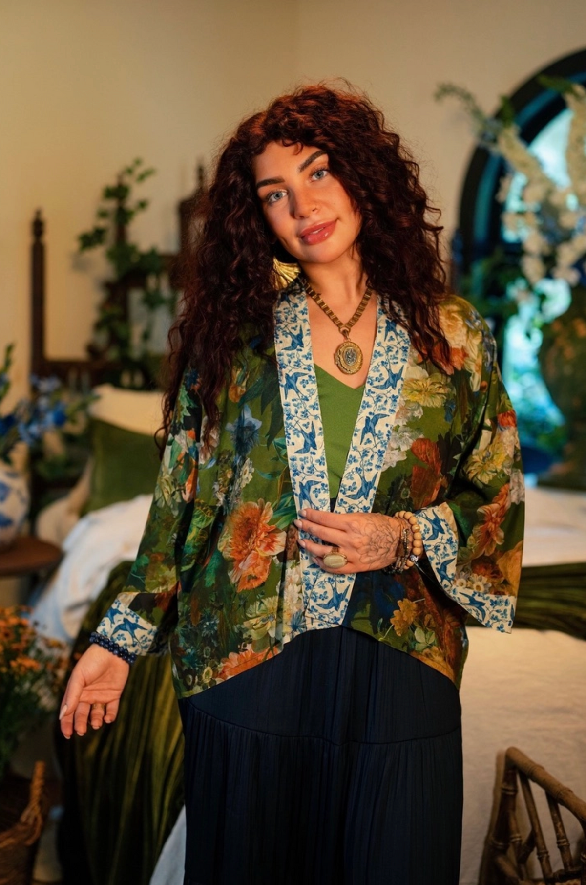 A woman with long, curly dark brown hair stands in a bedroom setting wearing a green floral print open-front jacket over a green top and a dark pleated skirt. The jacket features a blue and white patterned inner panel and cuffs | Bliss by Cori
