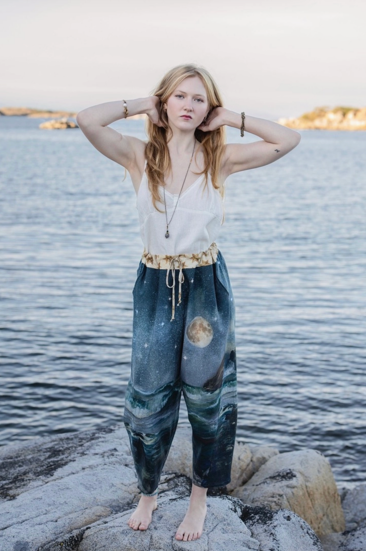 A woman stands barefoot on rocks by the water wearing loose harem-style pants with a galaxy print, a white sleeveless top, and a patterned belt | Bliss by Cori
