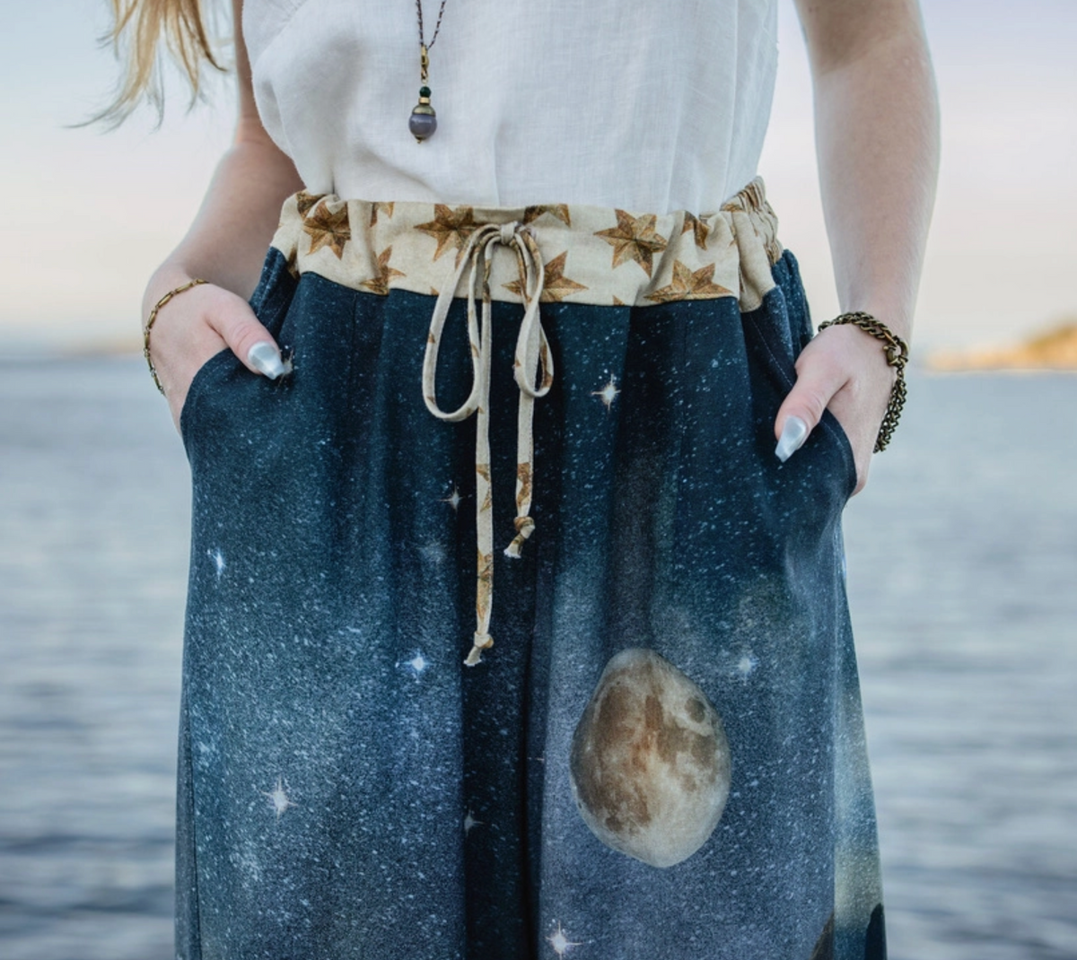 A person standing by the water wearing a white sleeveless top and dark blue galaxy-print wide-leg pants with a beige star-patterned drawstring waistband | Bliss by Cori