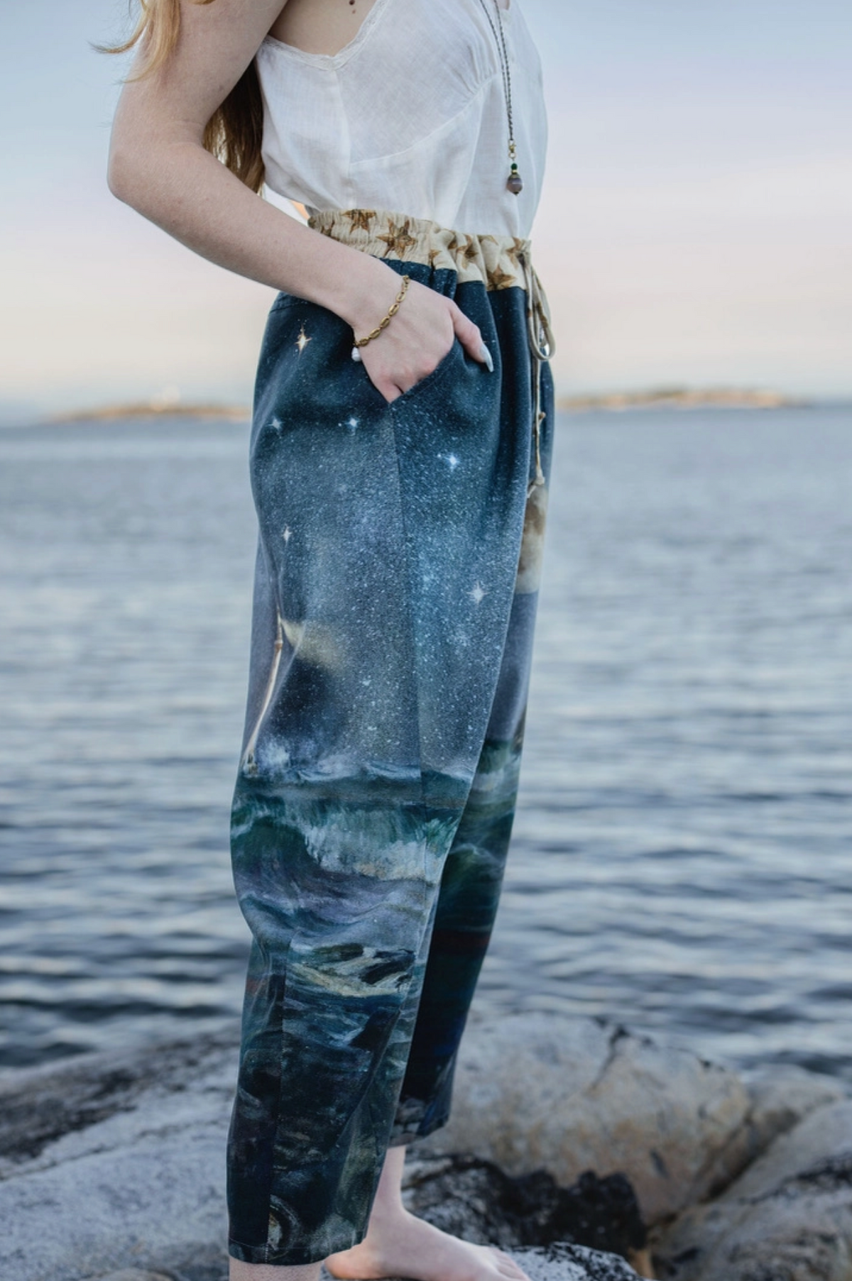 A person standing barefoot on rocks by the water, wearing loose dark blue pants with a galaxy and landscape print, a white sleeveless top, and a patterned belt | Bliss by Cori