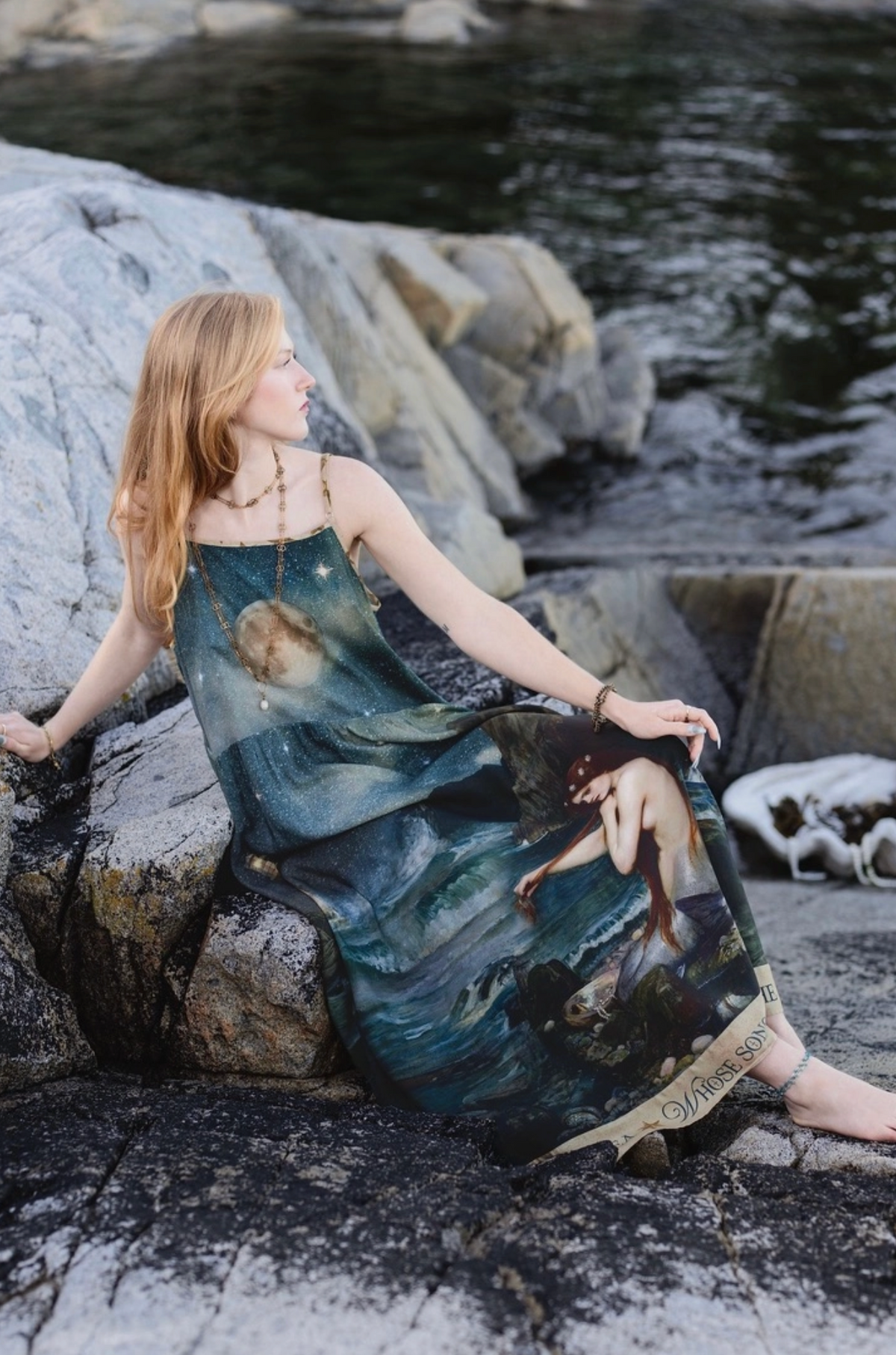 A woman sits on large rocks by the water wearing a long printed dress with a celestial, painterly design in dark teal, blue, and cream tones. A white dog is visible on the rocks beside her | Bliss by Cori