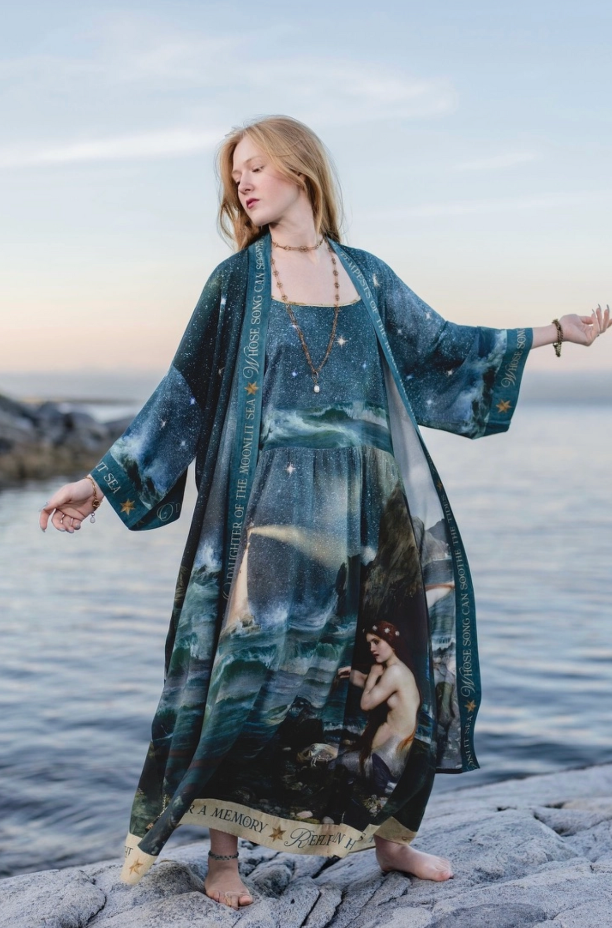 A woman stands barefoot on rocks by the water at sunset, wearing a long flowing sheer robe or kimono-style cover-up with a celestial and classical painting print in deep blue, teal, black, and beige tones | Bliss by Cori
