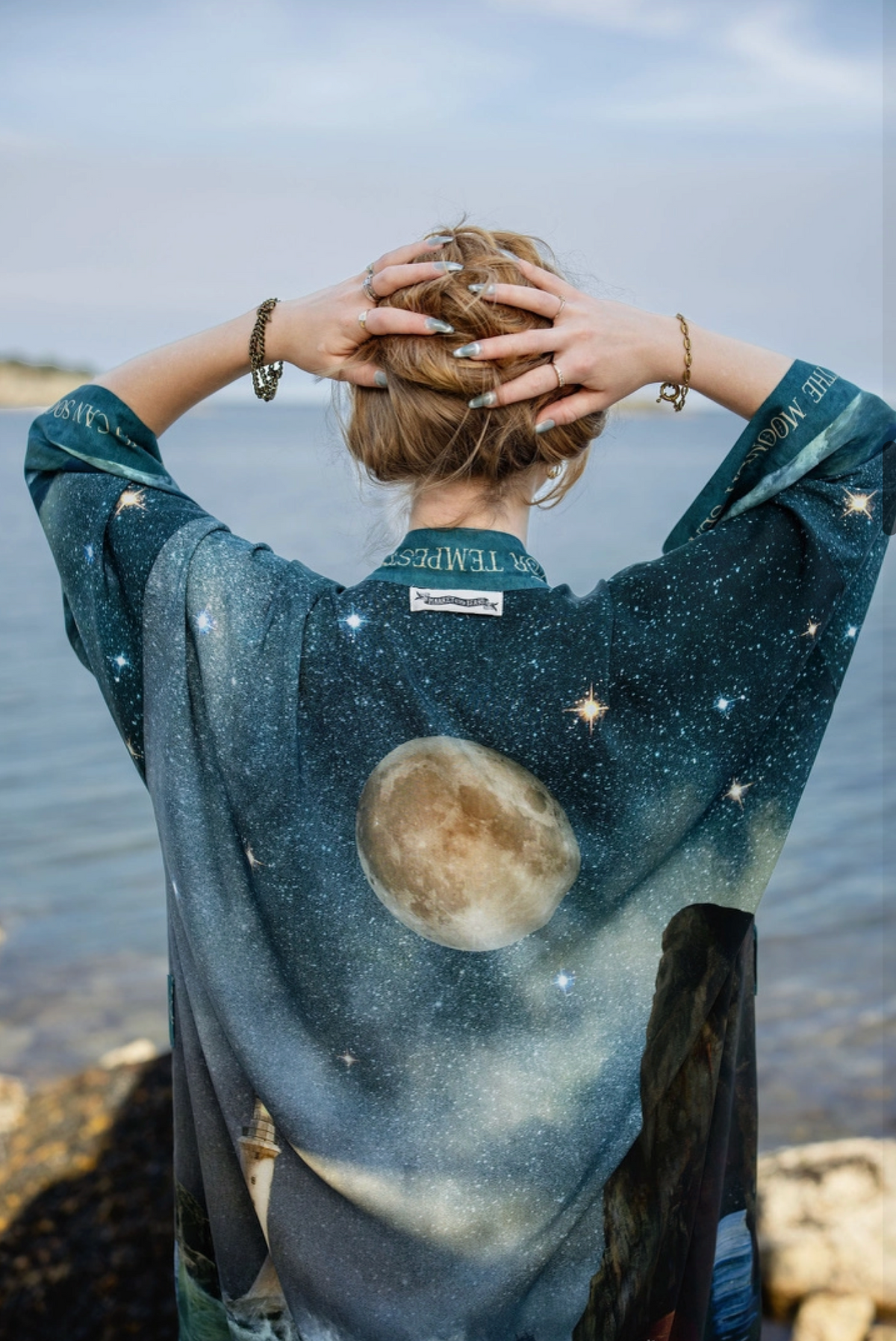 A woman stands with her back to the camera on a rocky shoreline, wearing a loose, wide-sleeved kimono-style top with a galaxy and moon print. The garment features a dark teal and black space design with white stars, a large moon graphic, and a scenic print along the lower edge | Bliss by Cori