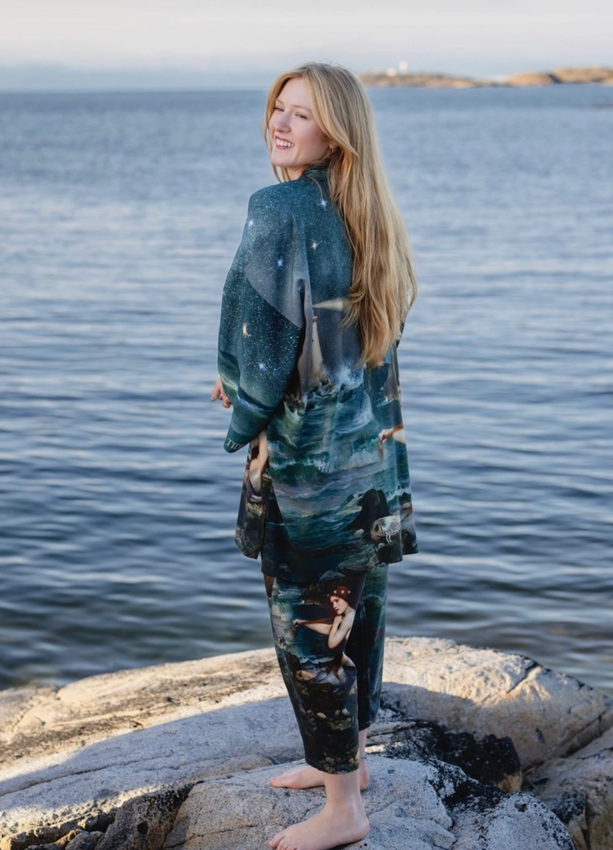 A woman with long blonde hair stands barefoot on rocks by the ocean, wearing a matching long-sleeve top and pants set with a dark blue and black celestial print featuring stars, moons, and a mermaid | Bliss by Cori