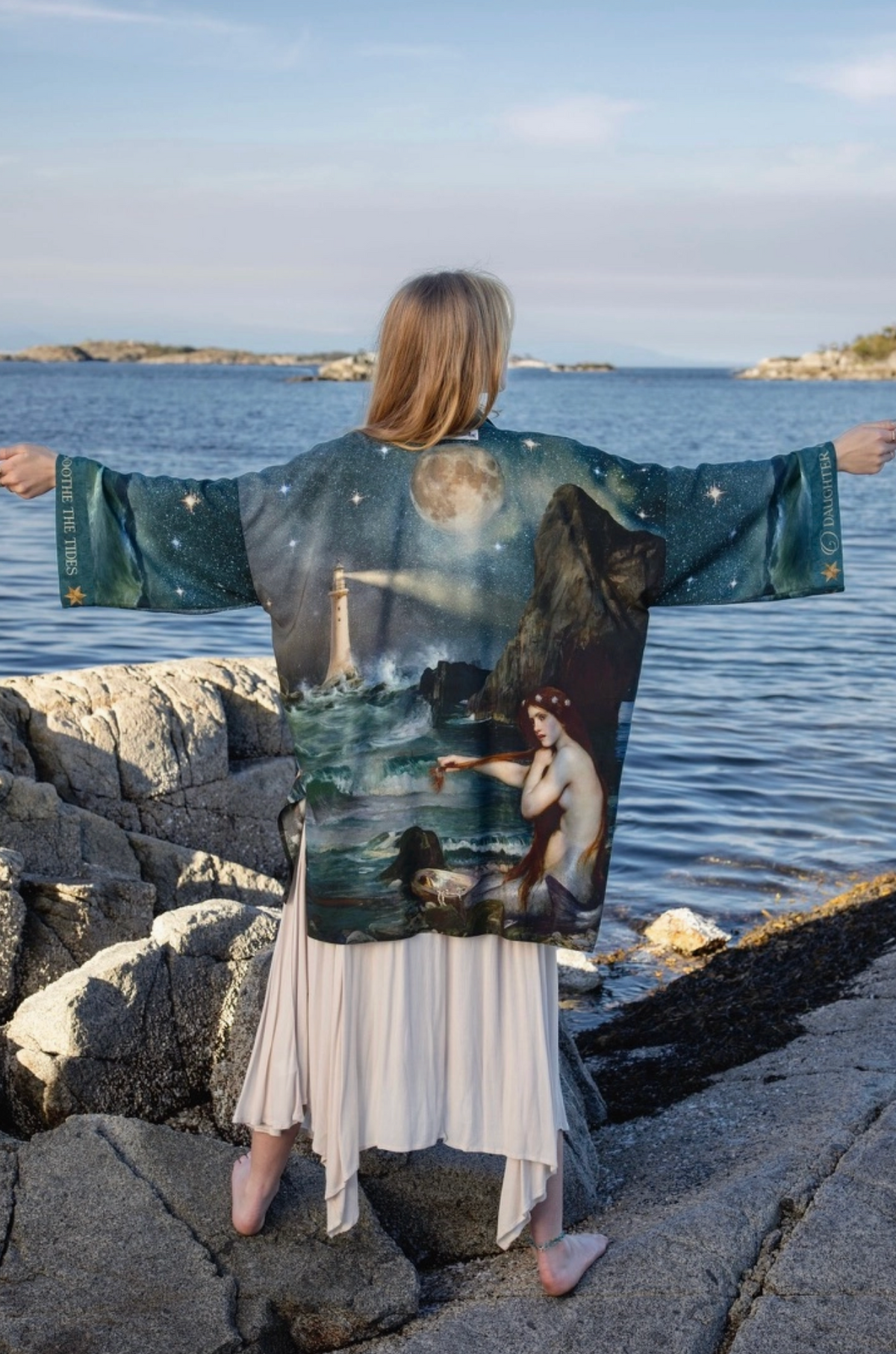 A woman stands on a rocky shoreline with her arms outstretched, wearing a wide-sleeved kimono-style robe printed with a fantasy scene of a mermaid, moon, and rocky landscape. The background shows water, rocks, and distant land under a blue sky | Bliss by Cori