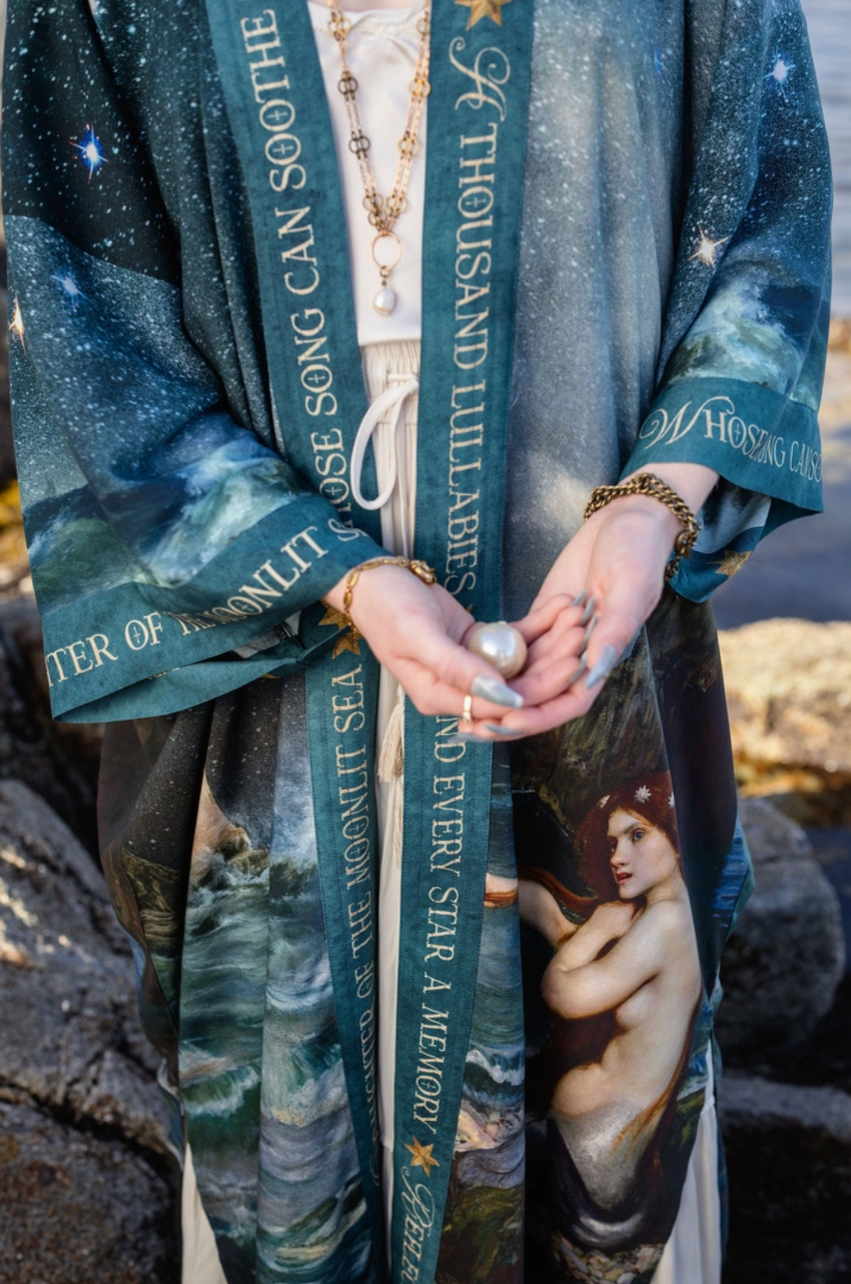 A person outdoors wearing a flowing printed robe or shawl with celestial and portrait artwork, draped over a white dress. The fabric features teal, blue, and dark tones with gold text and star motifs, and the scene includes rocks and natural light | Bliss by Cori