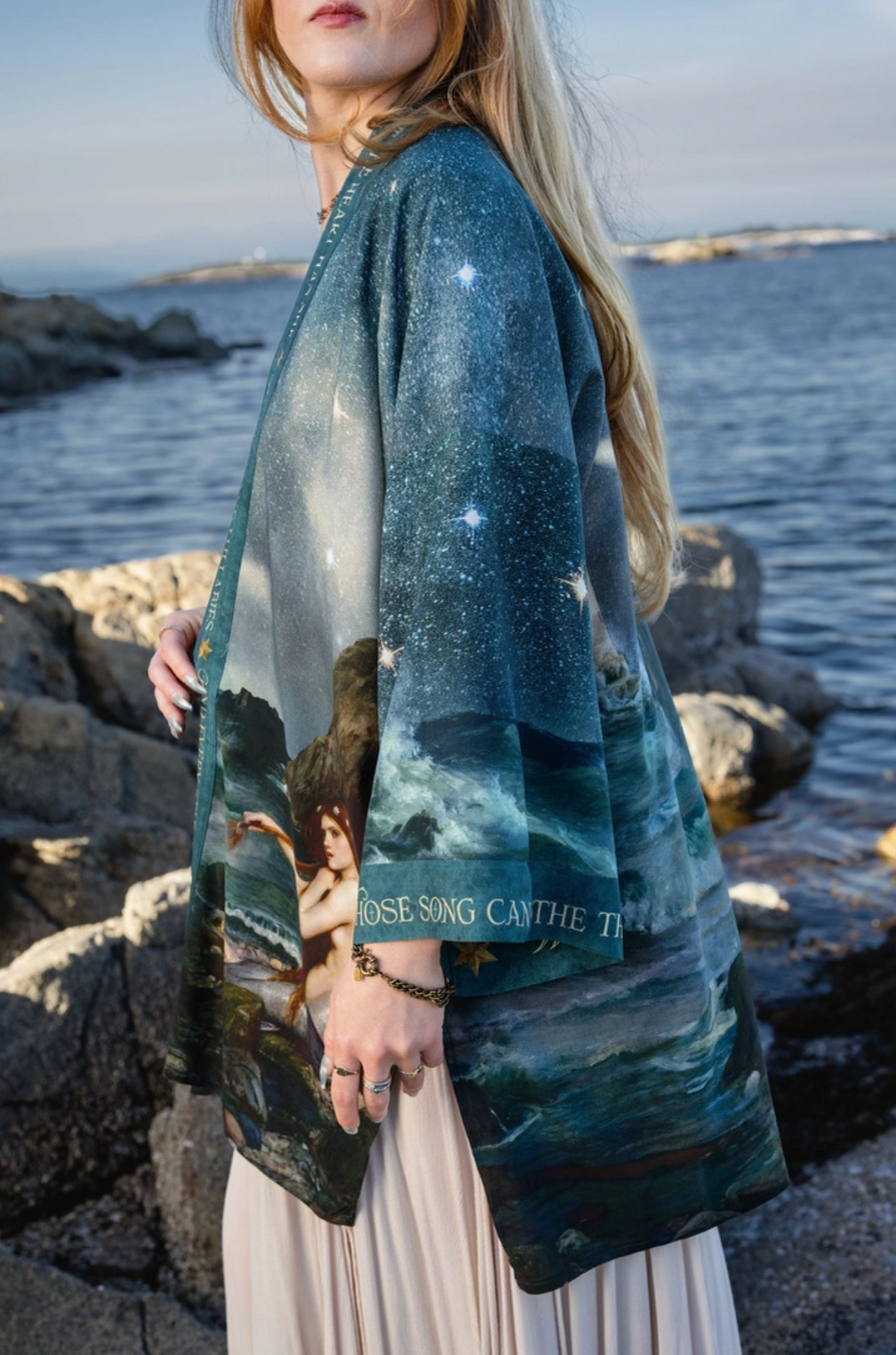 A woman standing on a rocky shoreline by the water, wearing a loose, open-front robe or kimono-style top with a celestial and scenic print in deep teal, blue, and white, paired with a light beige skirt | Bliss by Cori