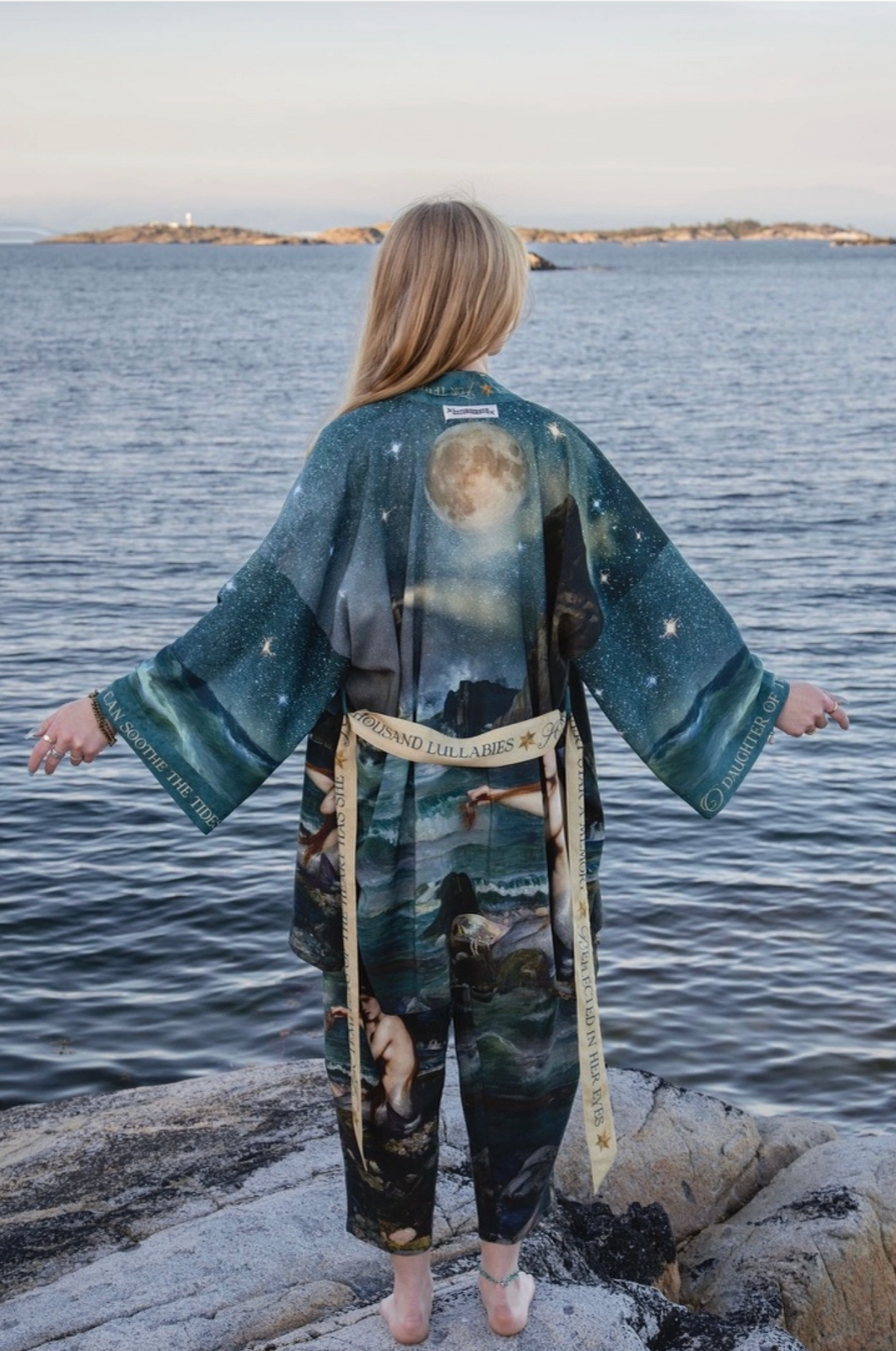A woman stands on rocks by the ocean with her back to the camera, wearing a long-sleeved kimono-style robe and matching pants in a dark teal and black celestial print with a beige belt | Bliss by Cori