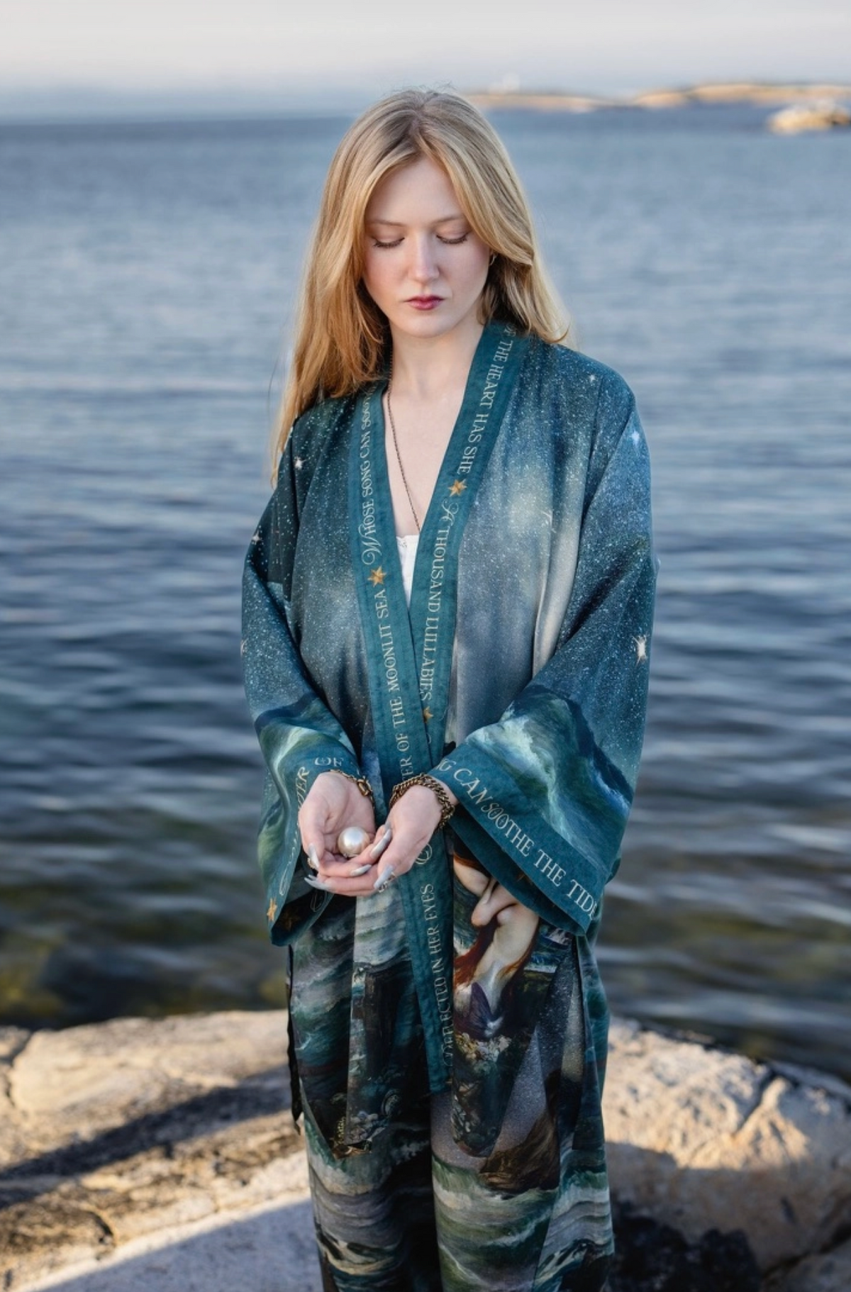 A woman stands on a rocky shoreline with the ocean behind her, wearing a long teal and blue printed robe | Bliss by Cori