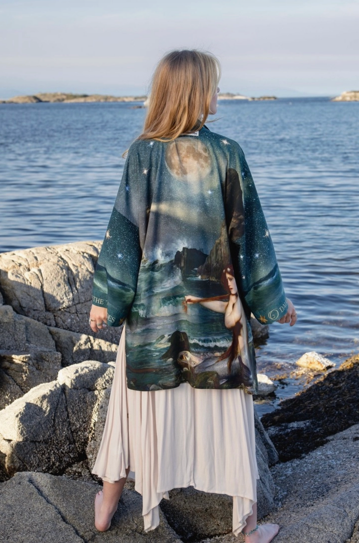 A woman stands on rocky shores facing the ocean, wearing a long-sleeved printed kimono-style jacket with a celestial and mermaid-themed design over a cream pleated maxi skirt | Bliss by Cori