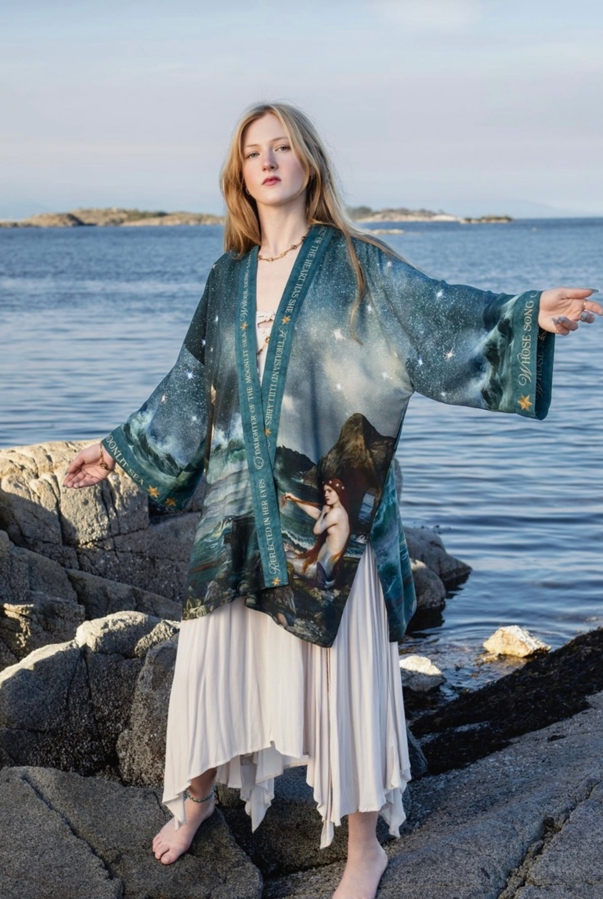 A woman stands barefoot on rocks by the ocean, wearing a long teal and blue printed kimono-style cardigan with a celestial and scenic print, layered over a white pleated dress | Bliss by Cori