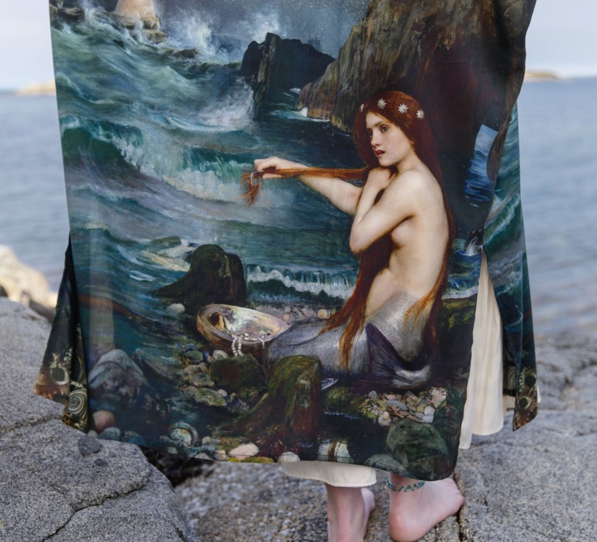 A draped fabric or wrap featuring a detailed print of a mermaid seated among rocks and waves, worn by a person standing outdoors on a rocky shoreline with water in the background | Bliss by Cori