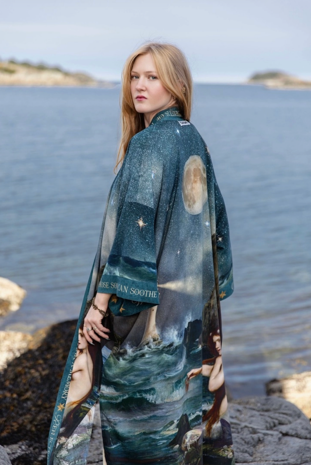 A woman stands on a rocky shoreline by the water, wearing a long printed wrap or shawl with a celestial and nautical design in deep blue, teal, and cream tones. The fabric includes moon and star motifs, a moon phase, and illustrated figures near the lower edge | Bliss by Cori