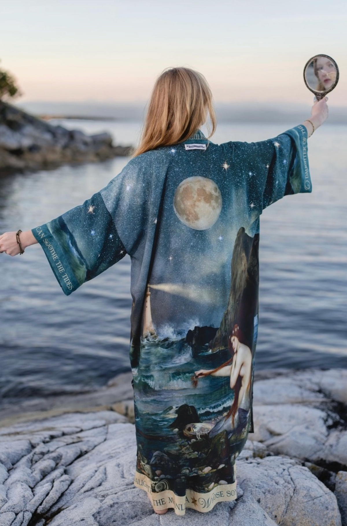A woman stands on rocks by the water, photographed from behind, wearing a long printed kimono-style robe with a celestial and mythic scene featuring a large moon, stars, sea, and a mermaid. She holds a small hand mirror in one hand | Bliss by Cori