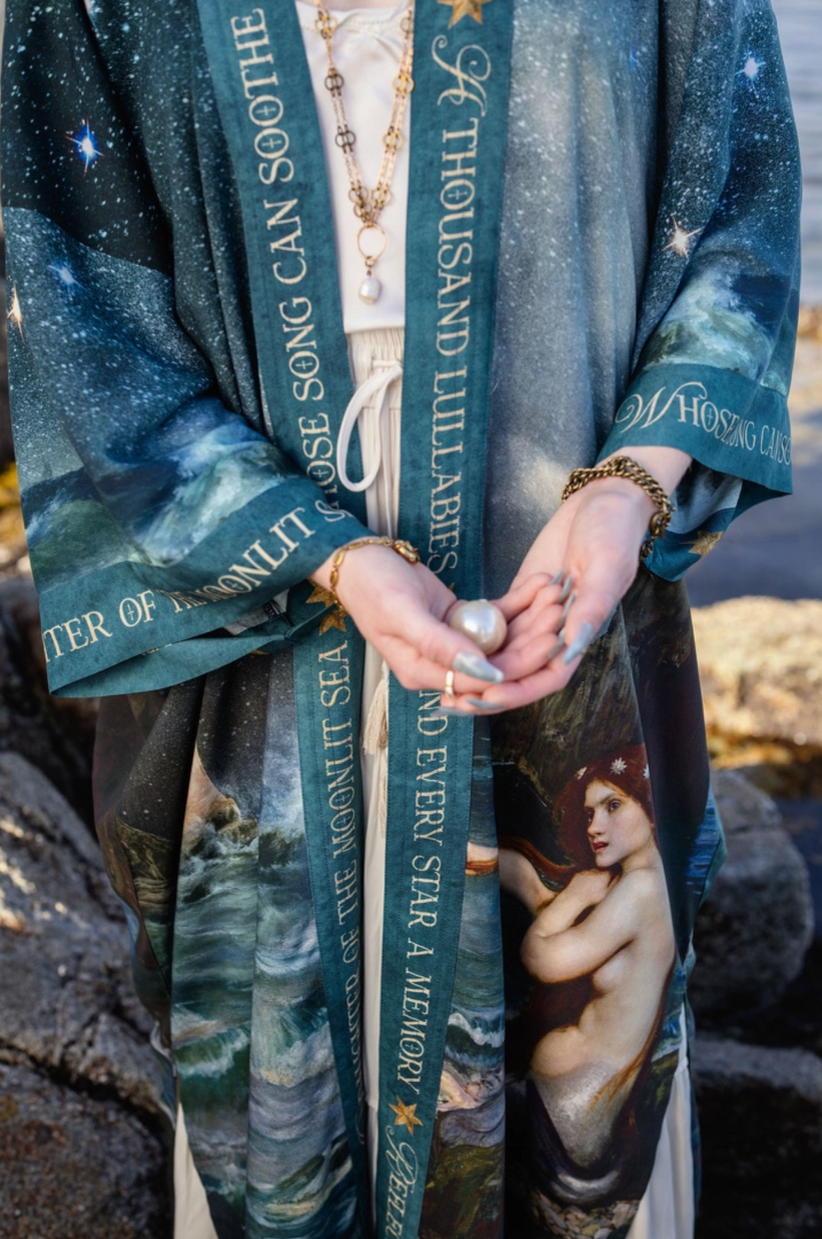 A close-up of a person outdoors wearing a long teal and navy celestial robe with printed starry sky and classical artwork panels, holding a small white stone in their hands | Bliss by Cori