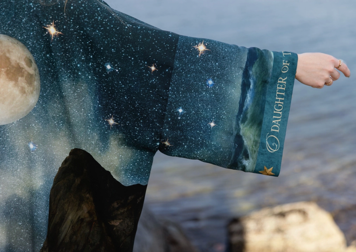 A person outdoors by the water wearing a long flowing robe or garment printed with a starry night sky, moon, and ocean scene. The sleeve cuff reads 'DAUGHTER OF THE" | Bliss by Cori