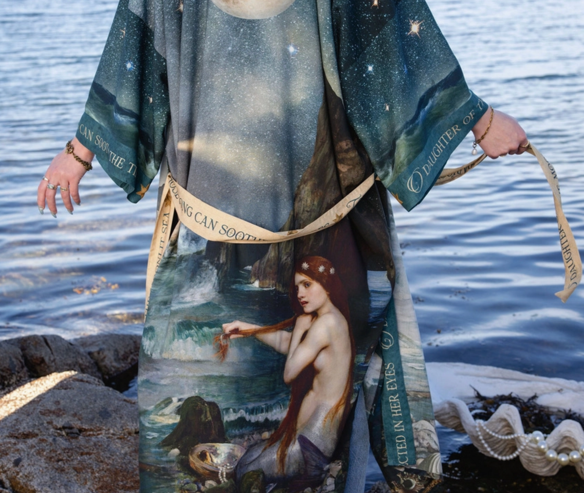 A person wearing a long printed robe or kimono with a mermaid and starry seascape design, standing at the water's edge and holding a printed ribbon. The garment has a dark blue and teal celestial print with a large mermaid illustration on the front and text running along the sleeves and lower hem | Bliss by Cori
