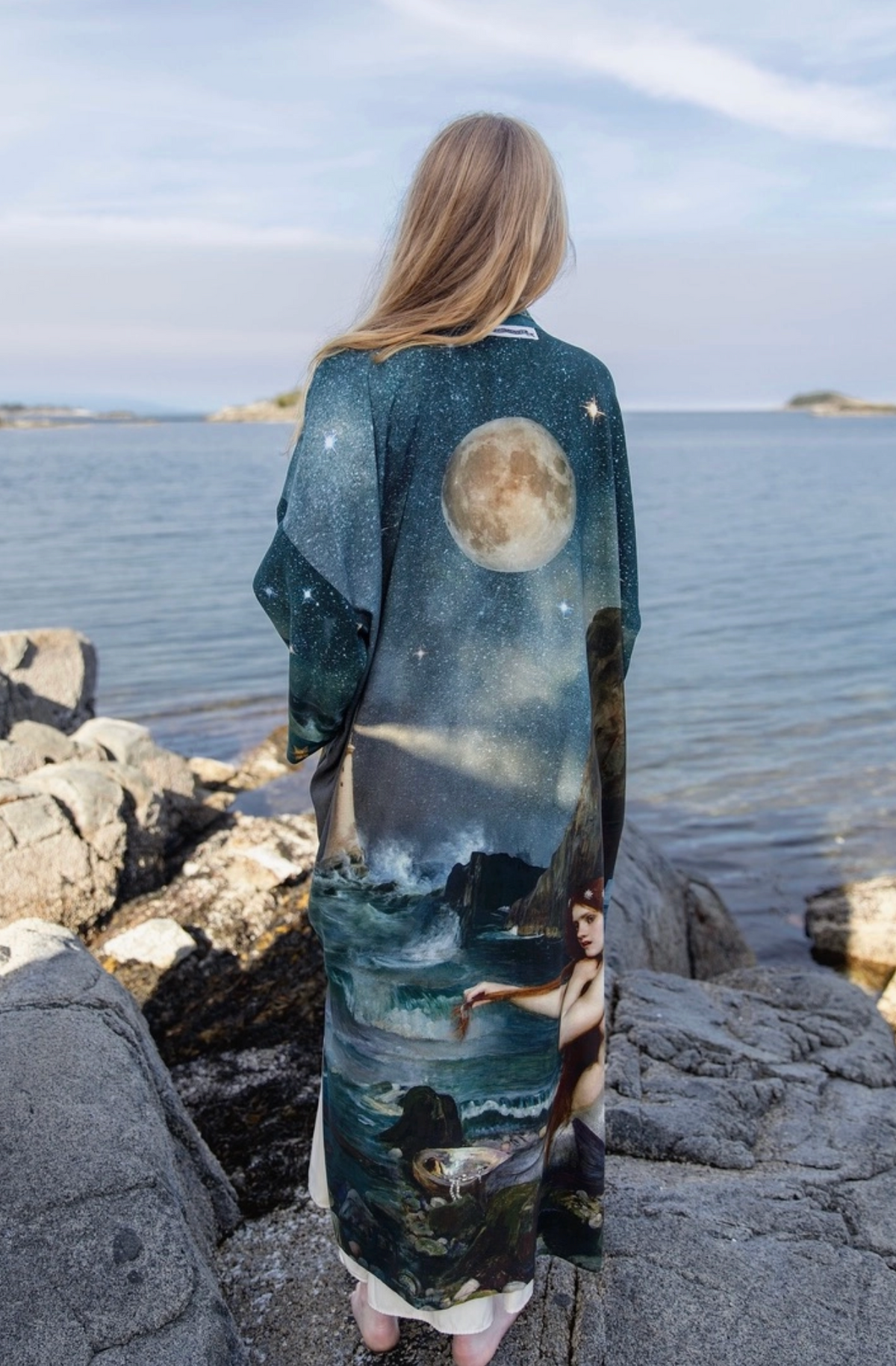 Model wearing the full-length Siren duster on shoreline rocks with the moon artwork visible on the back | Bliss by Cori