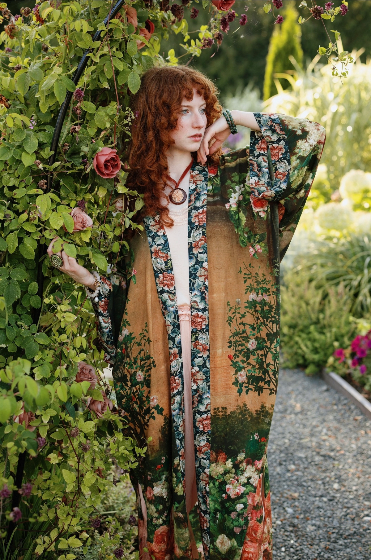 A woman with red hair stands in a garden wearing a long, open-front floral robe over a white top. The robe features a tan base with green, pink, and red botanical prints, and is styled with a long wooden pendant necklace | Bliss by Cori