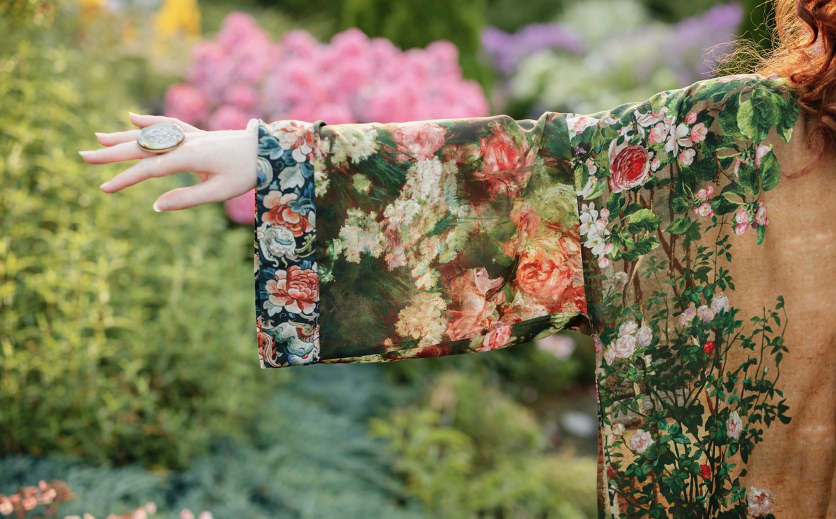 A person wearing a long-sleeved floral robe or kimono-style garment, photographed outdoors in a garden with their arm extended. The garment features a rich, painterly floral pattern with pink, red, green, and cream tones, and a solid tan sleeve cuff | Bliss by Cori