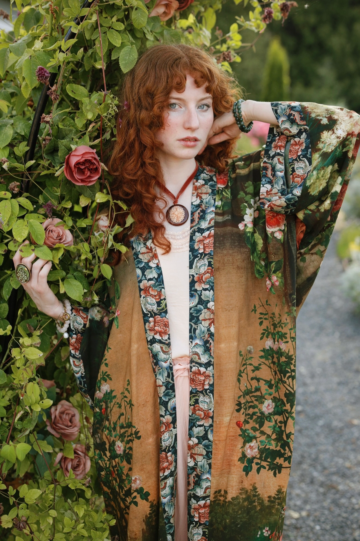 A woman with curly red hair stands outdoors beside a leafy rose wall, wearing a long open-front kimono-style robe with a tan base and floral print, over a white top. The robe features dark green and orange botanical patterns with a central vertical scenic panel | Bliss by Cori