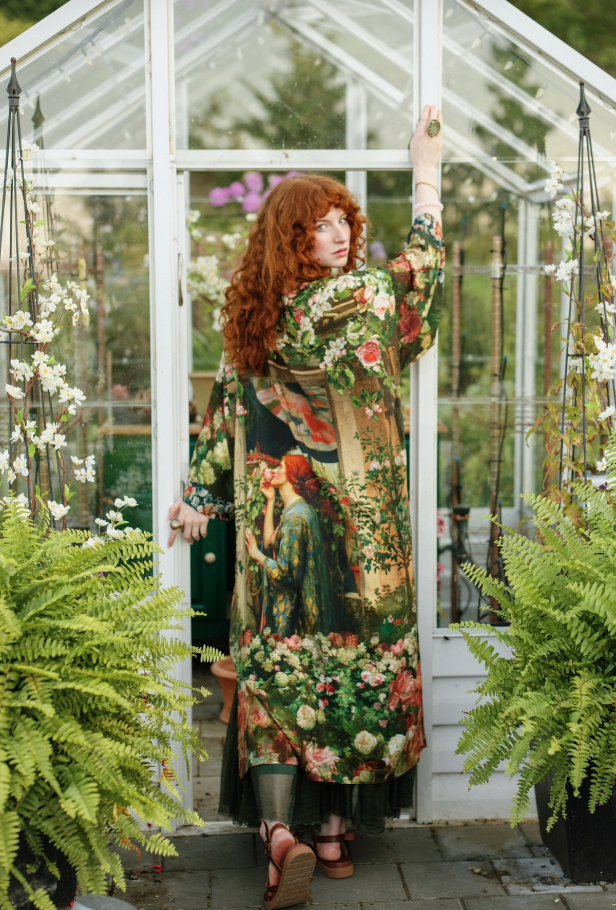A woman with long red hair stands in a greenhouse doorway wearing a long-sleeved, floor-length floral and scenic print kimono-style robe. The robe features a detailed vintage-style illustration of a woman in a landscape with prominent green foliage, red flowers, and a warm beige background | Bliss by Cori