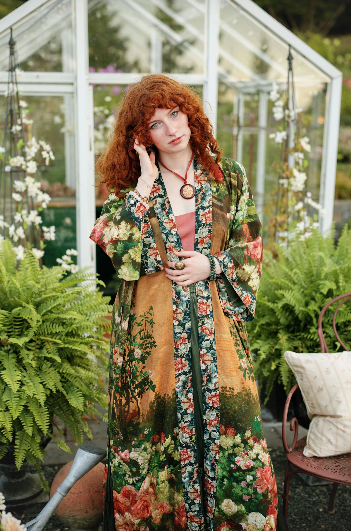 A woman with vibrant red hair stands in a greenhouse wearing a long-sleeved, open-front floral robe over a dusty pink top. The robe features a detailed botanical print with green foliage, orange-brown backgrounds, and red and pink flowers | Bliss by Cori