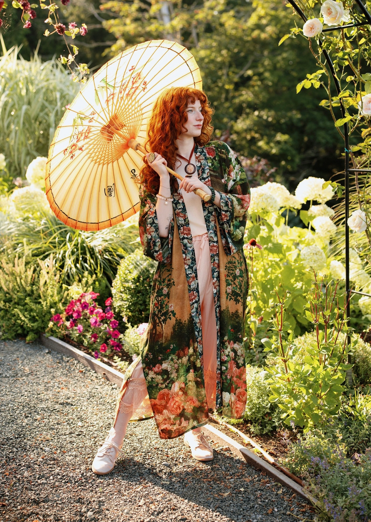 A woman stands in a garden holding a large beige paper umbrella. She is wearing a long, flowing floral robe with a pink lining and white shoes | Bliss by Cori
