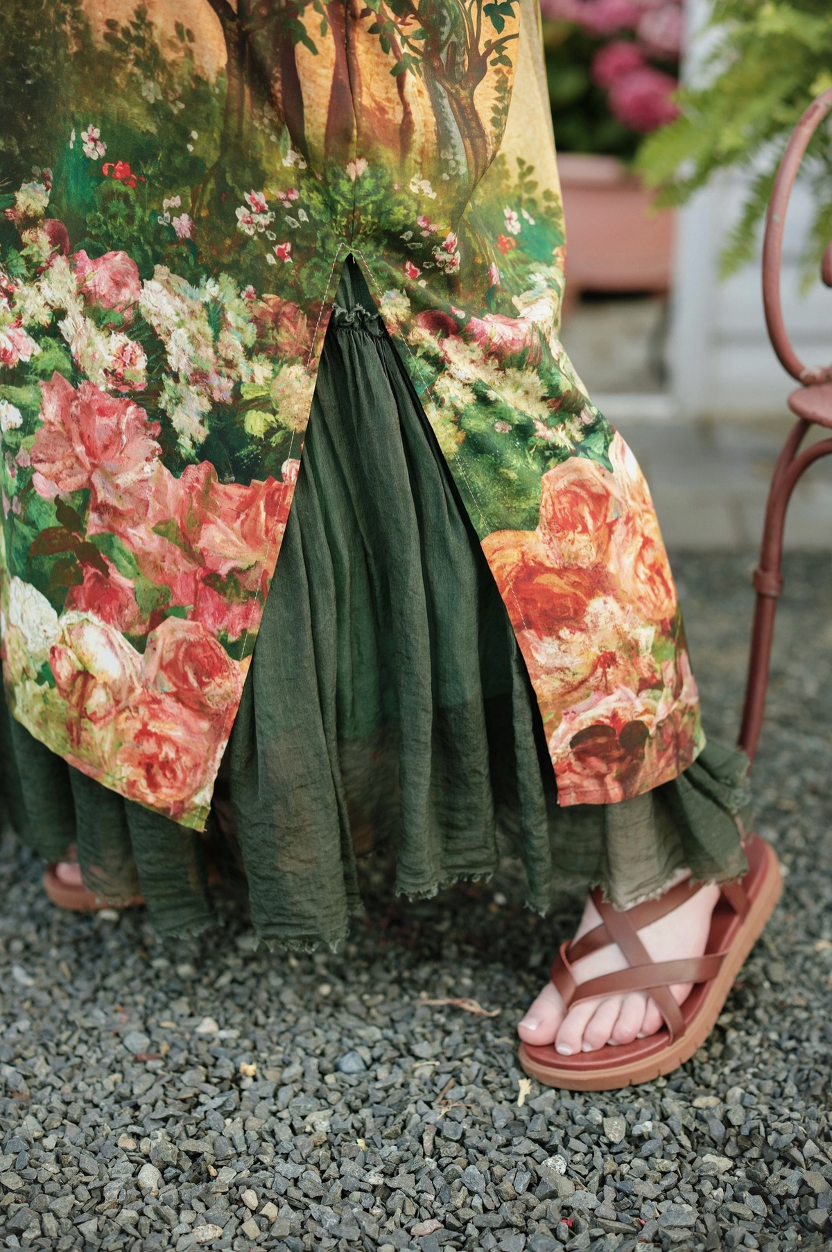 A close-up of a person's feet wearing brown leather flat sandals and a dark green skirt with a large floral print overlay, standing on a gravel surface | Bliss by Cori
