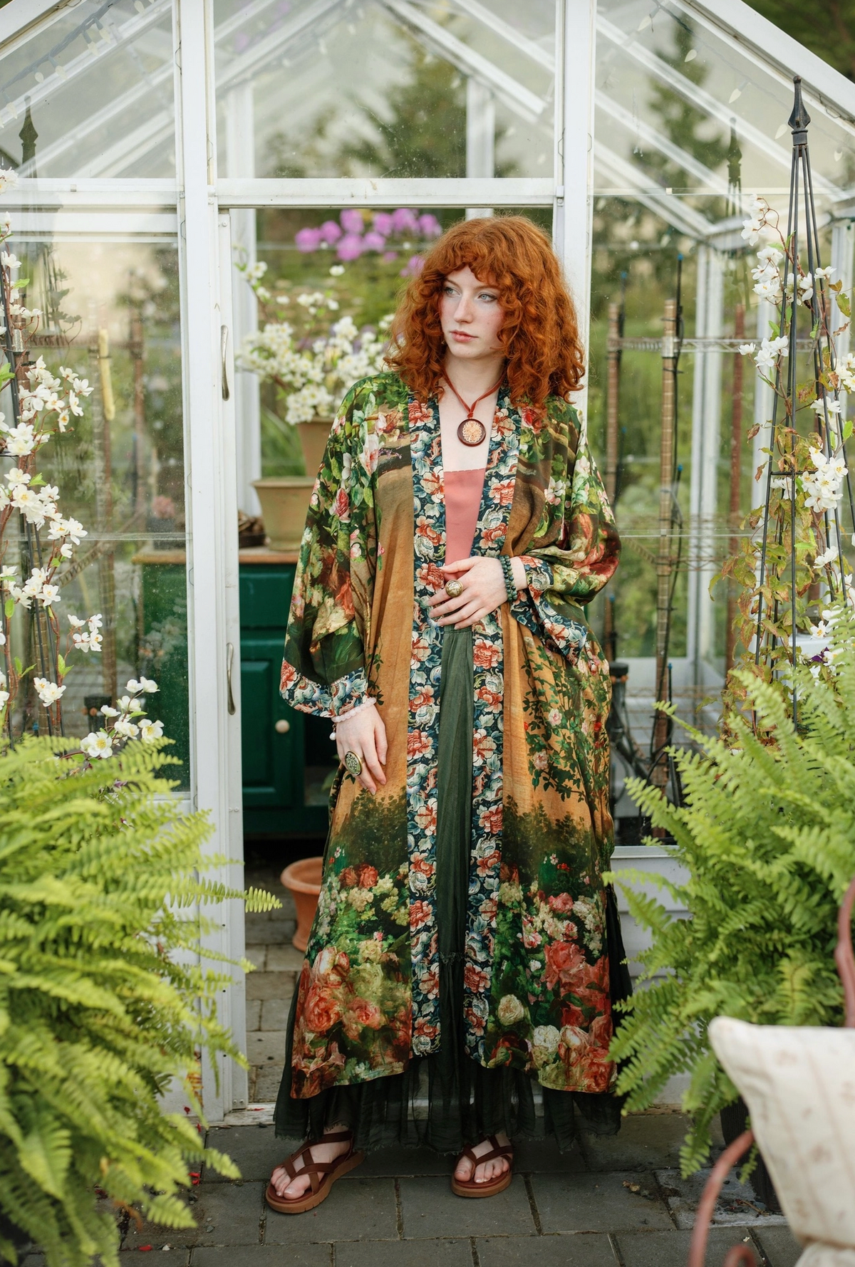 A woman with curly red hair stands in a greenhouse wearing a long bohemian floral kimono jacket over a dark green maxi dress. The jacket features a multicolor floral print with green, orange, and brown tones, and she is wearing brown leather sandals | Bliss by Cori