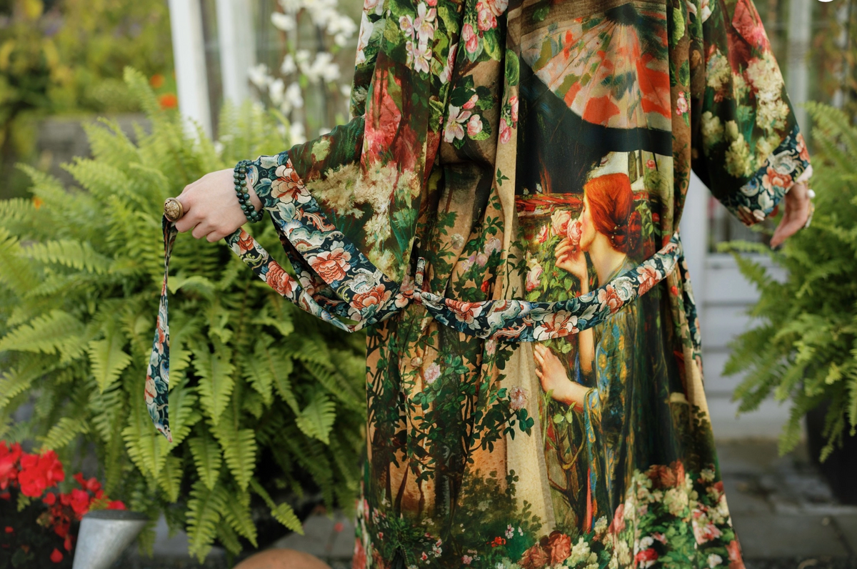A person wearing a long-sleeved, knee-length floral and scenic print robe or kimono-style dress with a matching tie belt, standing in a garden setting with green ferns in the background | Bliss by Cori