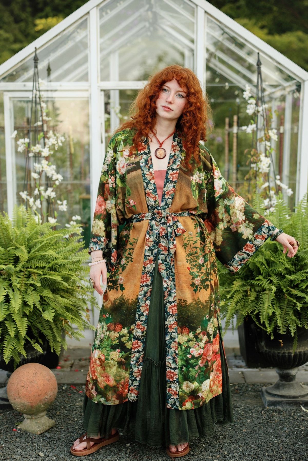 A woman with red hair stands in front of a glass greenhouse wearing a long-sleeved, belted floral kimono-style jacket over a dark green dress. She is surrounded by potted ferns on a gravel path | Bliss by Cori