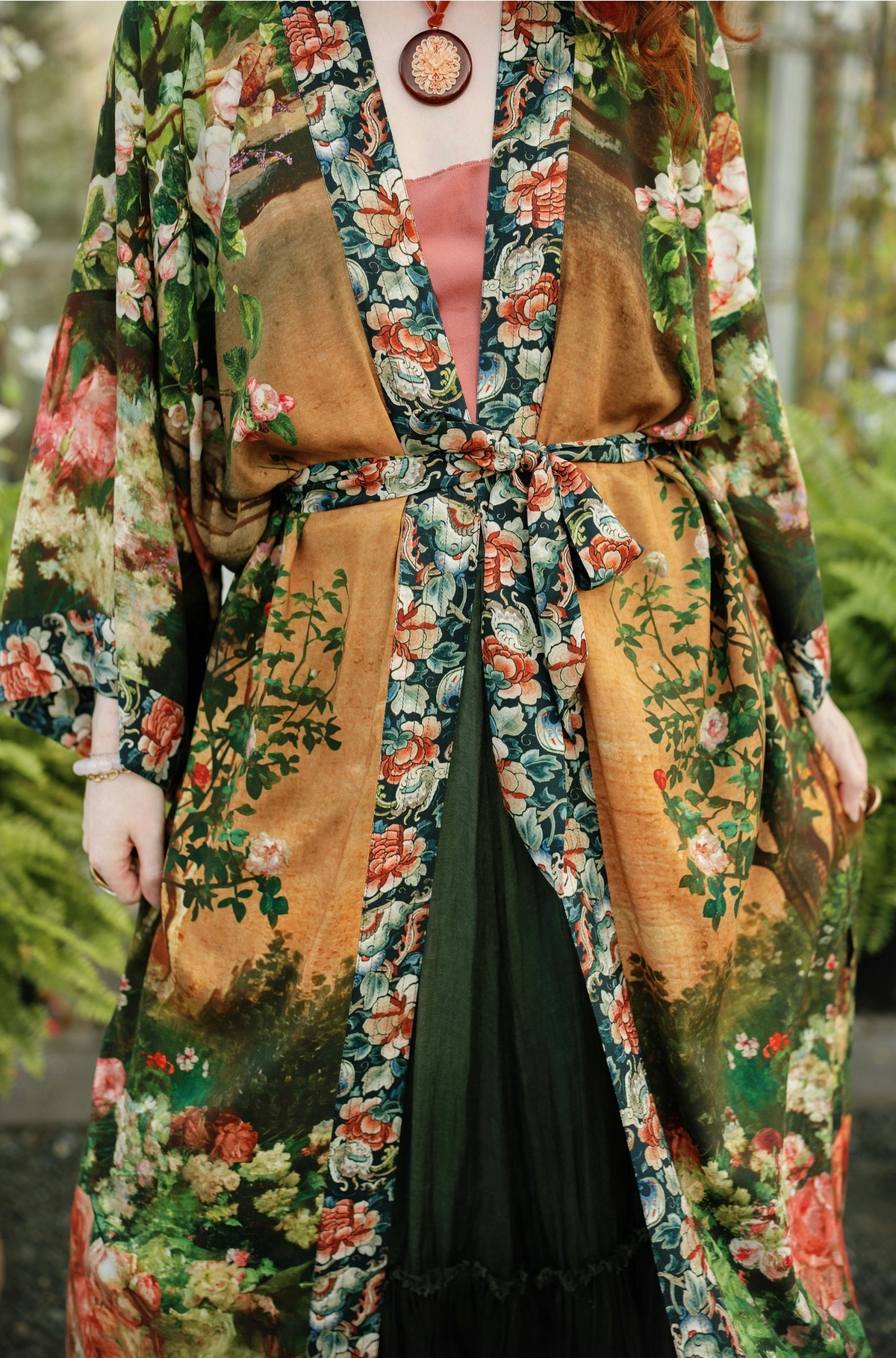 A person wearing a long, wide-sleeved floral robe or kimono-style outer layer over a dark green dress, photographed outdoors with blurred greenery in the background | Bliss by Cori
