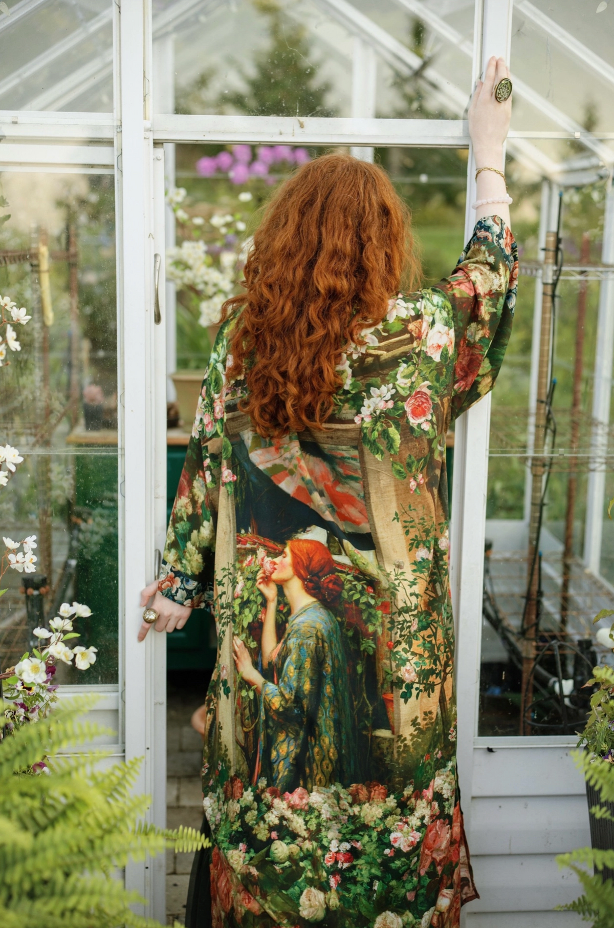 A woman with long curly red hair stands inside a glass greenhouse, viewed from behind as she wears a long-sleeved floral robe with an illustrated print of a red-haired woman in a garden | Bliss by Cori