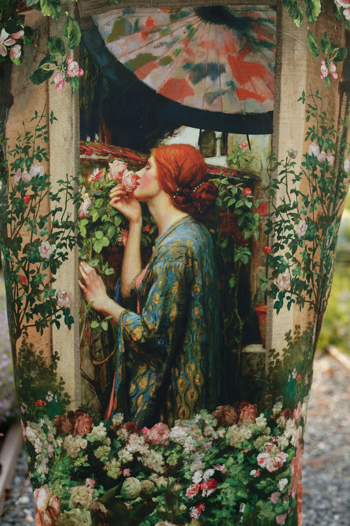 A large decorative planter or vase featuring a detailed classical-style painting of a red-haired woman in a blue patterned dress among roses, photographed outdoors against a garden background | Bliss by Cori