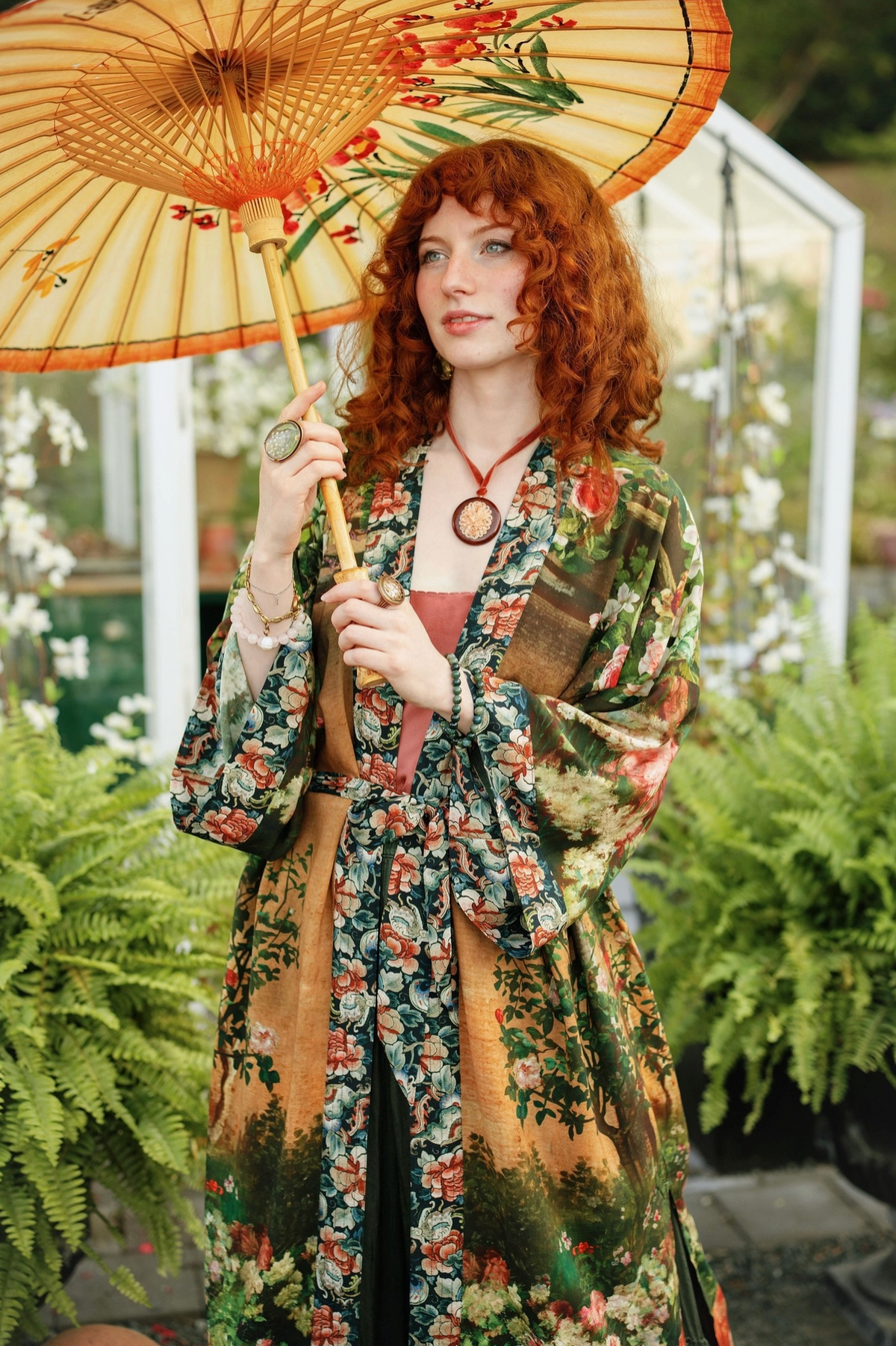 A woman with curly red hair stands in a greenhouse holding a decorative paper umbrella. She is wearing a long-sleeved floral kimono robe with a brown patchwork panel on the front, layered over a pink top | Bliss by Cori