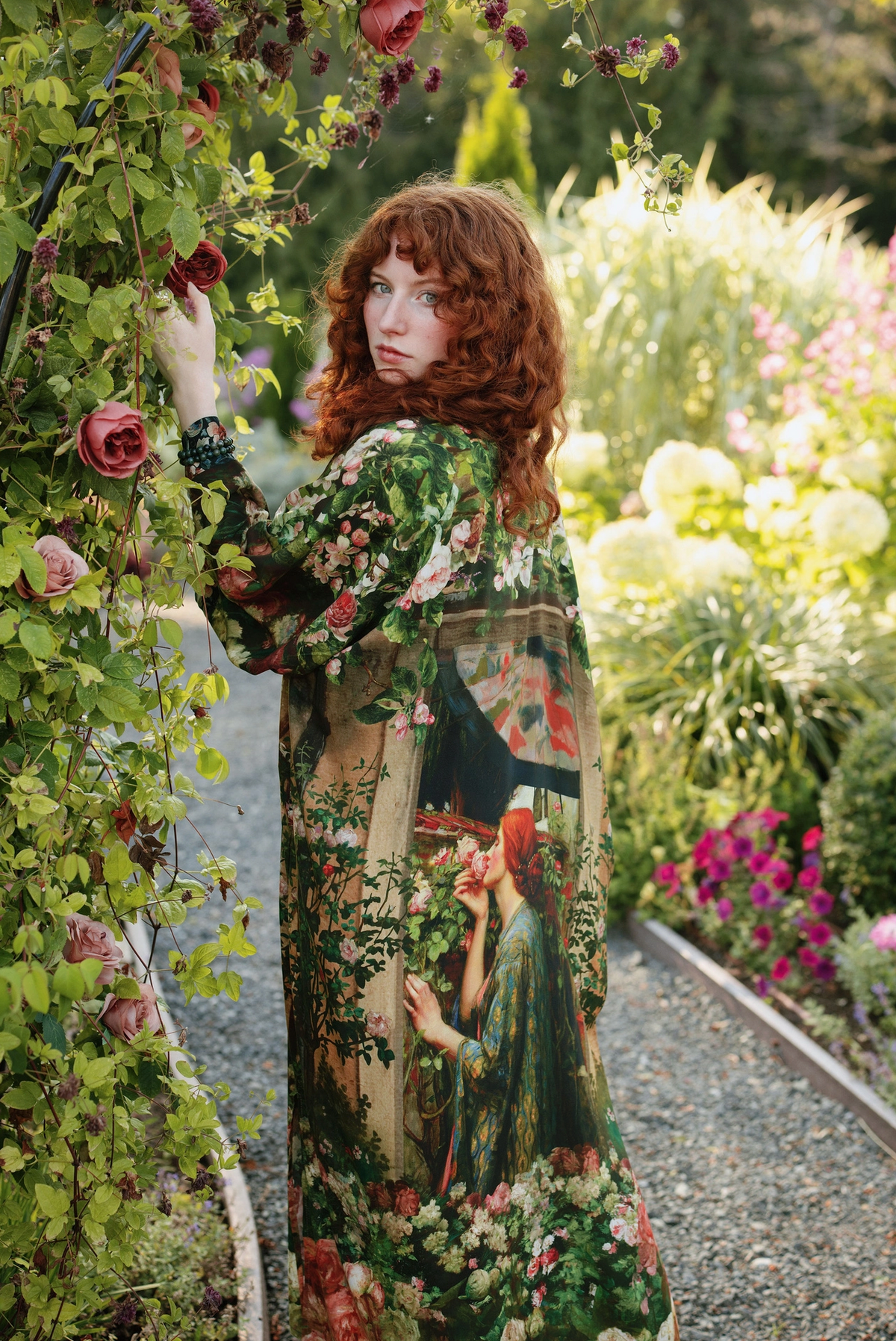 A woman with curly red hair stands in a lush garden wearing a long-sleeved maxi dress with a vintage-style botanical and portrait print. She is looking back over her shoulder while standing next to a rose-covered trellis | Bliss by Cori