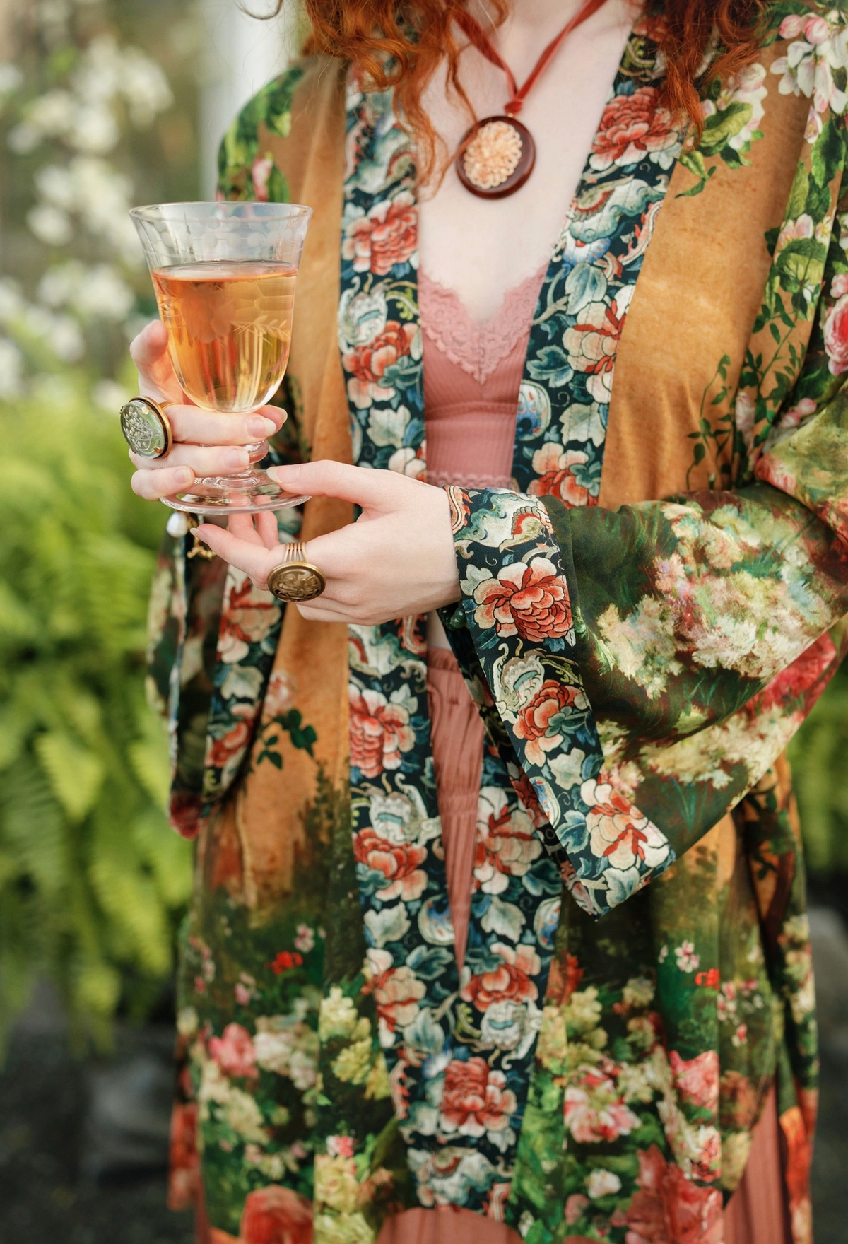 A close-up lifestyle shot of a person wearing a bohemian floral robe over a pink lace top, holding a glass of iced tea outdoors with green foliage in the background | Bliss by Cori