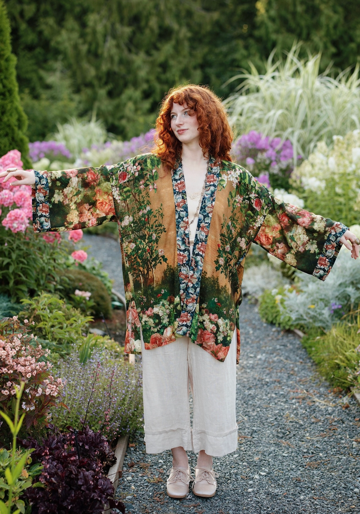 A woman with red hair stands on a gravel garden path wearing a floral kimono-style cardigan with wide sleeves, cream wide-leg pants, and beige shoes. The background is a lush green garden with pink and purple flowers | Bliss by Cori