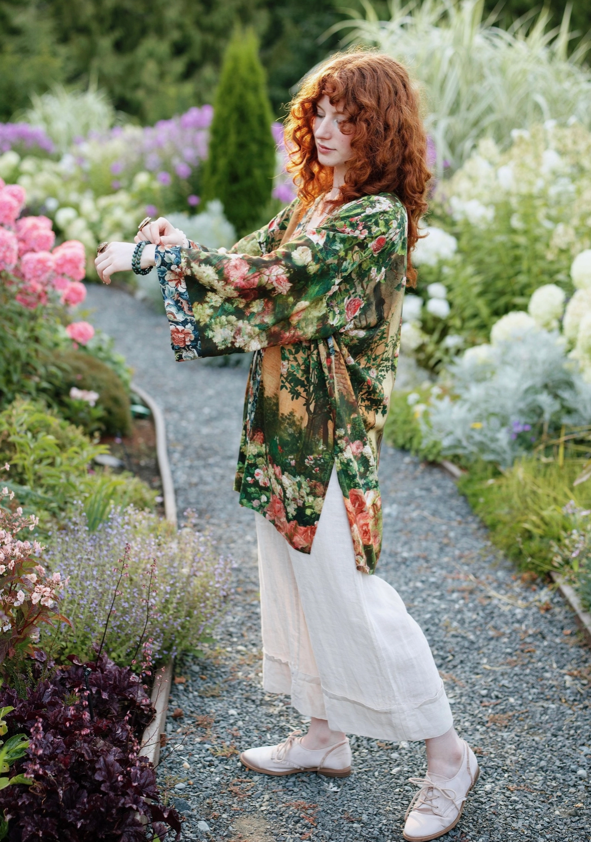 A woman with curly red hair stands in a garden wearing a loose floral kimono-style top with wide sleeves, a white skirt, and light pink shoes. She is looking down while adjusting a bracelet on her wrist | Bliss by Cori