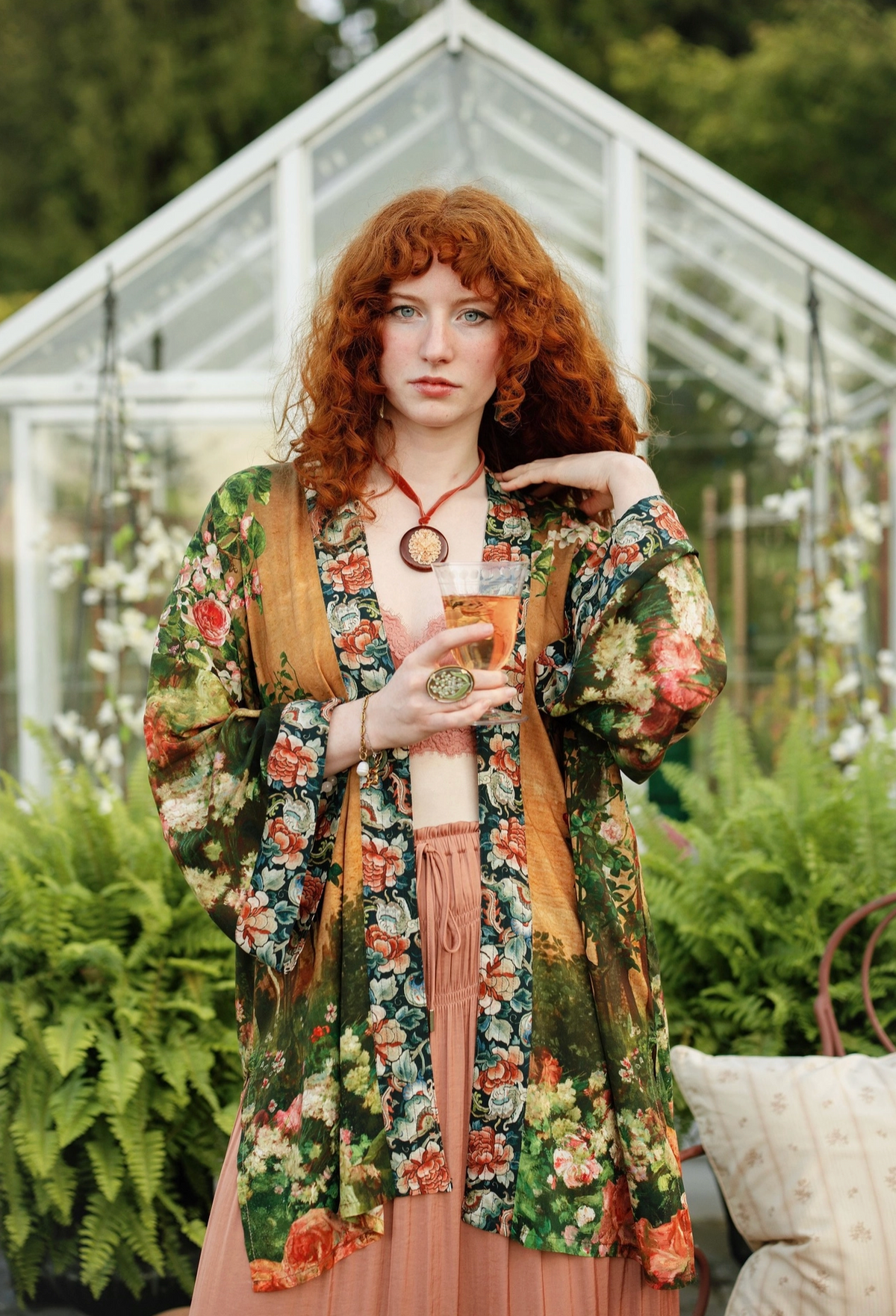 A woman with curly red hair stands in front of a greenhouse wearing a green floral cardigan over a pink dress, holding a glass of iced coffee | Bliss by Cori