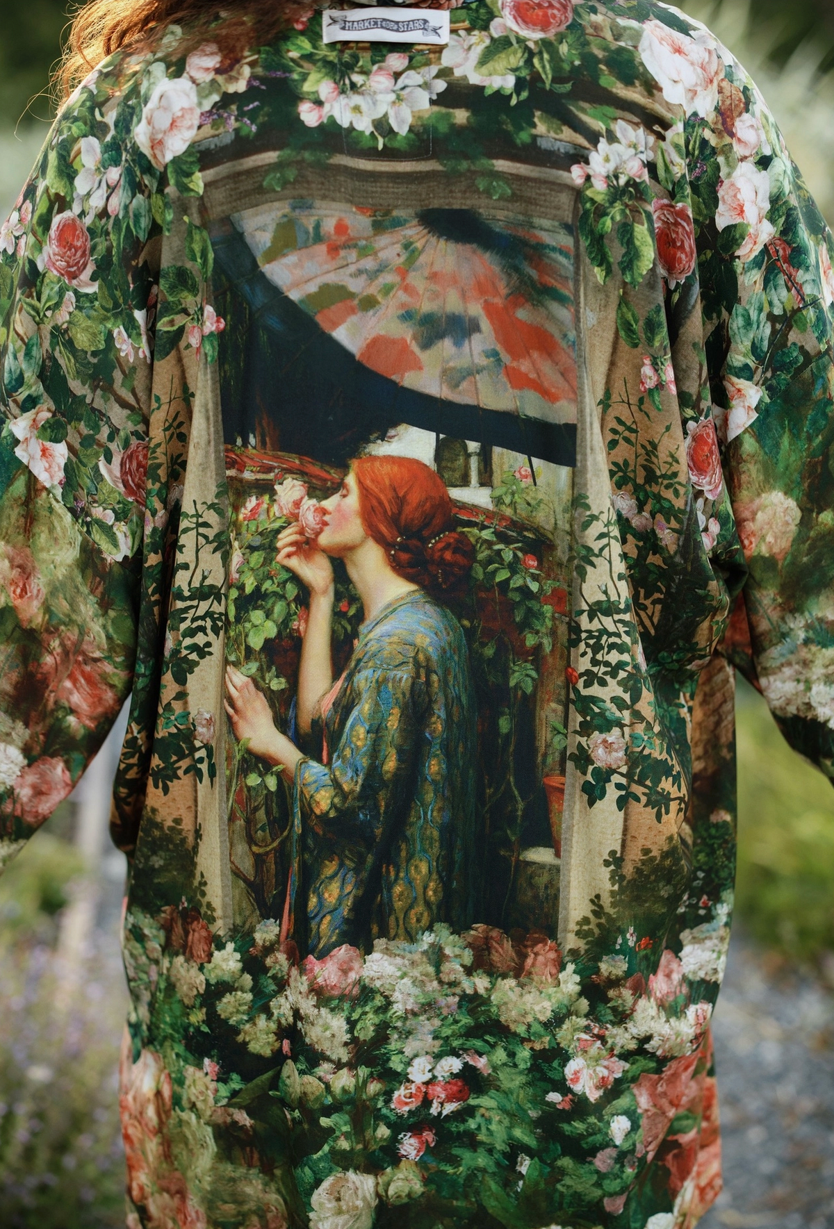 A close-up of a floral robe or kimono-style garment worn outdoors, featuring a large vintage-style painting print on the back showing a woman with red hair under an umbrella, surrounded by roses and foliage | Bliss By Cori