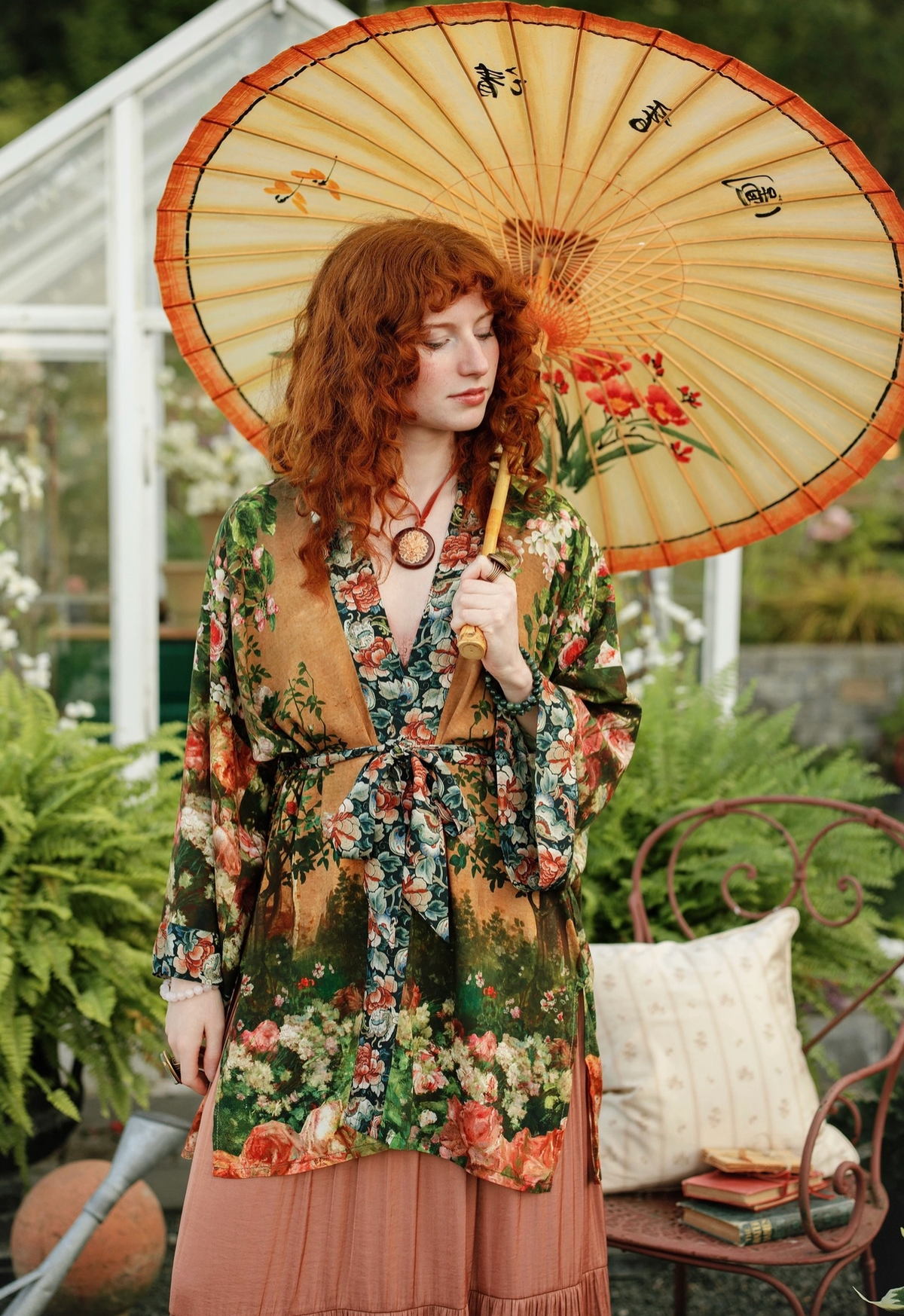 A woman with red hair stands in a garden holding a large beige paper parasol with orange trim and floral print. She wears a long-sleeve floral robe with a tan collar and a dusty pink skirt | Bliss by Cori