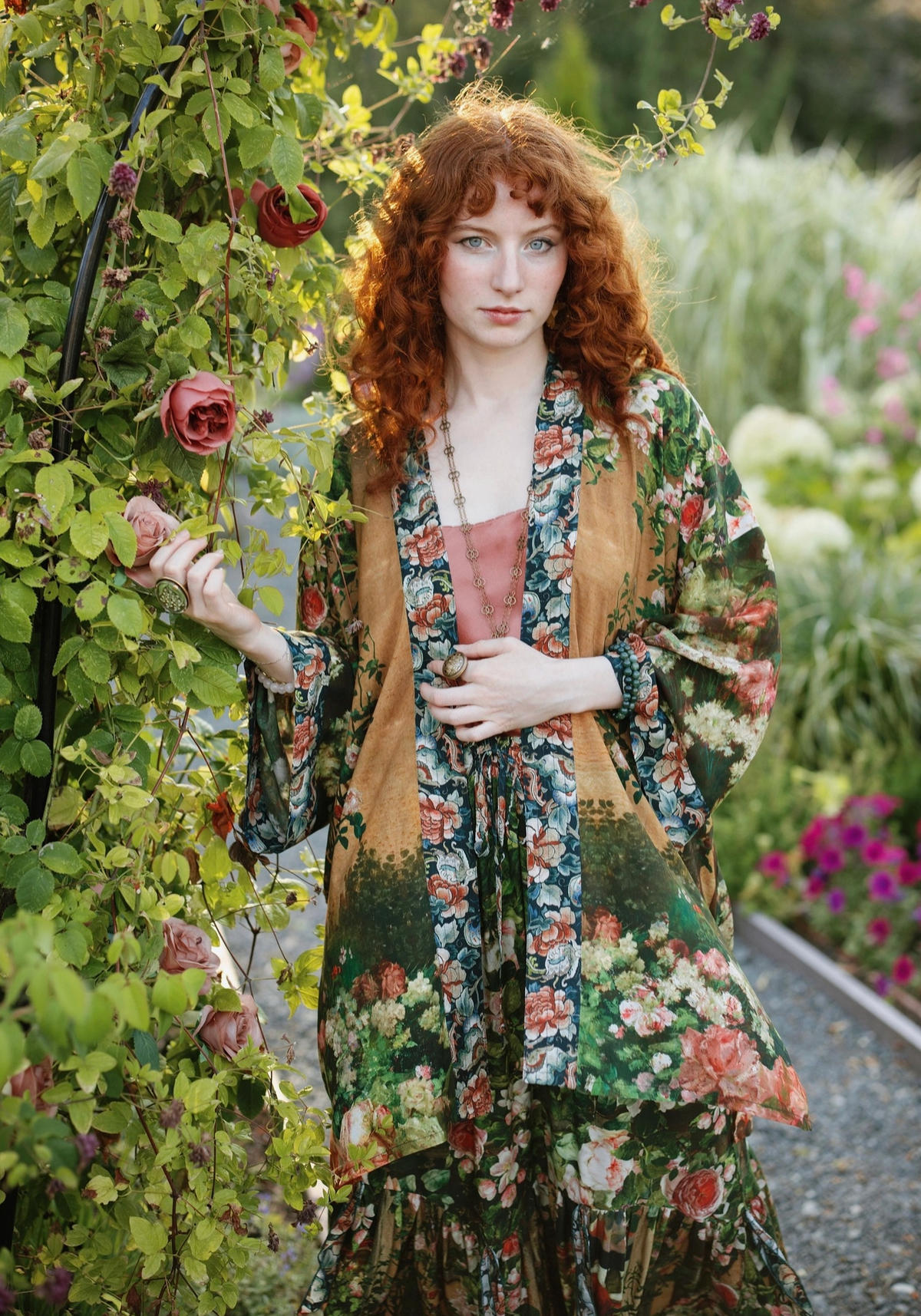 A woman with long red hair stands in a garden wearing a loose, long-sleeve floral robe over a dusty pink top. The robe features a multicolor floral pattern with green, pink, and brown tones | Bliss By Cori