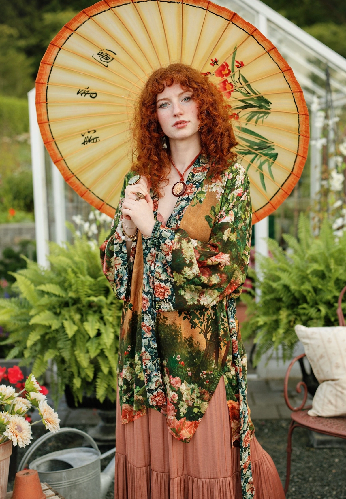A woman with long red hair stands in a greenhouse holding a large beige paper umbrella decorated with green leaves and red flowers. She is wearing a green floral kimono-style top over a dusty pink pleated skirt | Bliss by Cori
