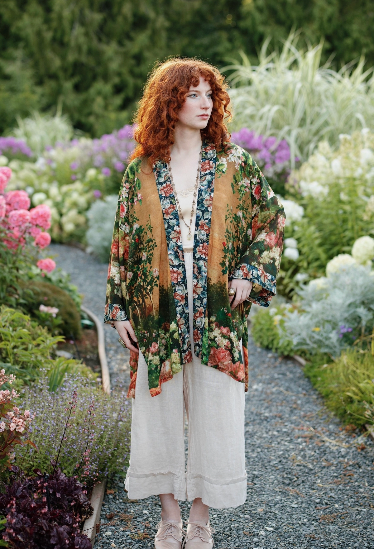 A woman with vibrant red hair stands on a gravel path in a lush, colorful garden, wearing a sheer floral kimono-style cardigan over a white linen dress | Bliss by Cori