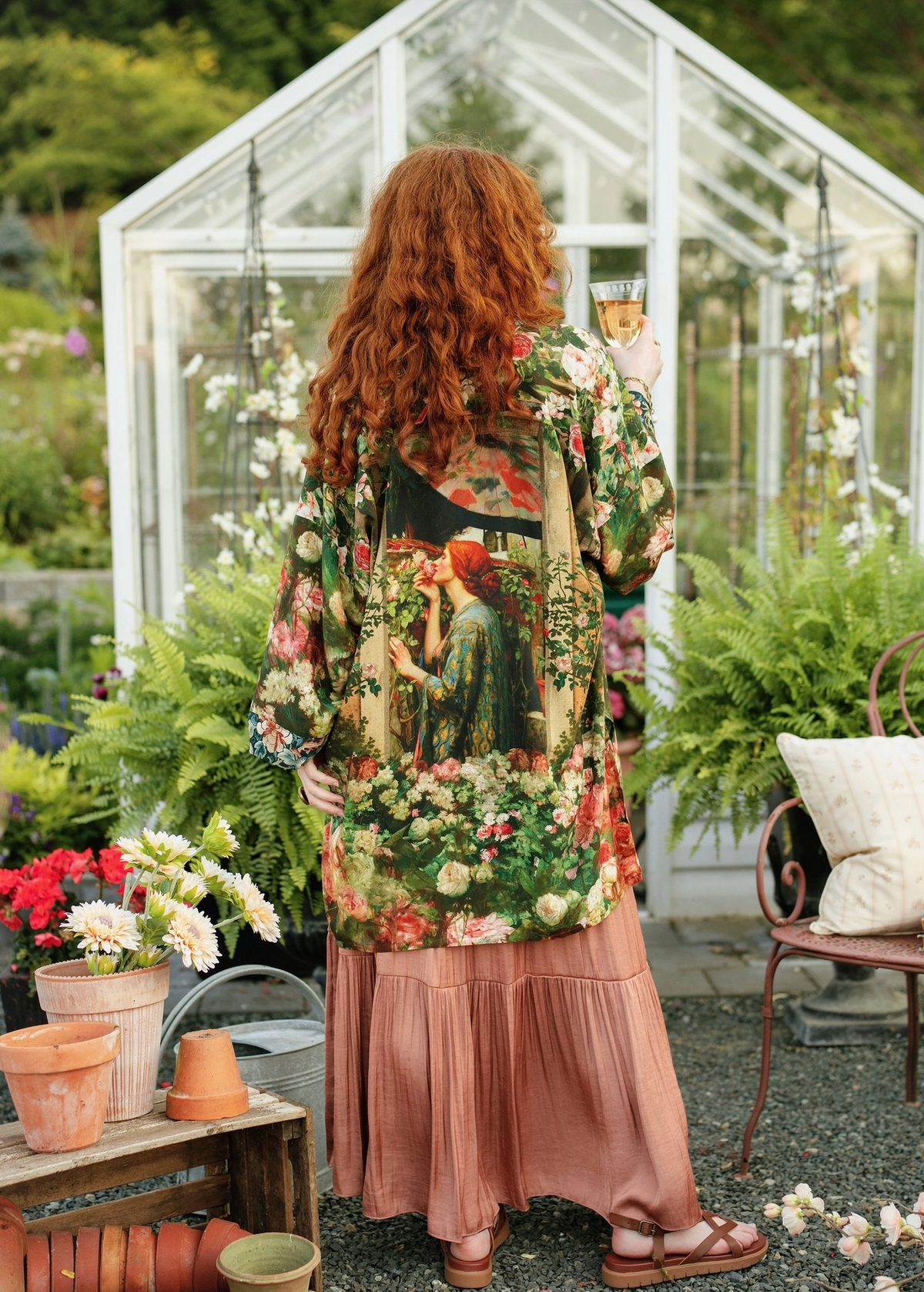 A woman with long red hair stands in a garden greenhouse setting, viewed from behind. She is wearing a long-sleeve button-up shirt with a detailed floral and botanical print featuring green foliage, red flowers, and a woman in a red dress, paired with a dusty rose pleated maxi skirt and brown sandals | Bliss by Cori