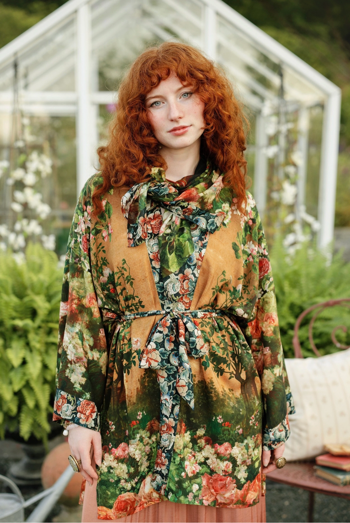 A woman with curly red hair wearing a long-sleeved, tie-neck floral robe in a greenhouse garden setting | Bliss by Cori